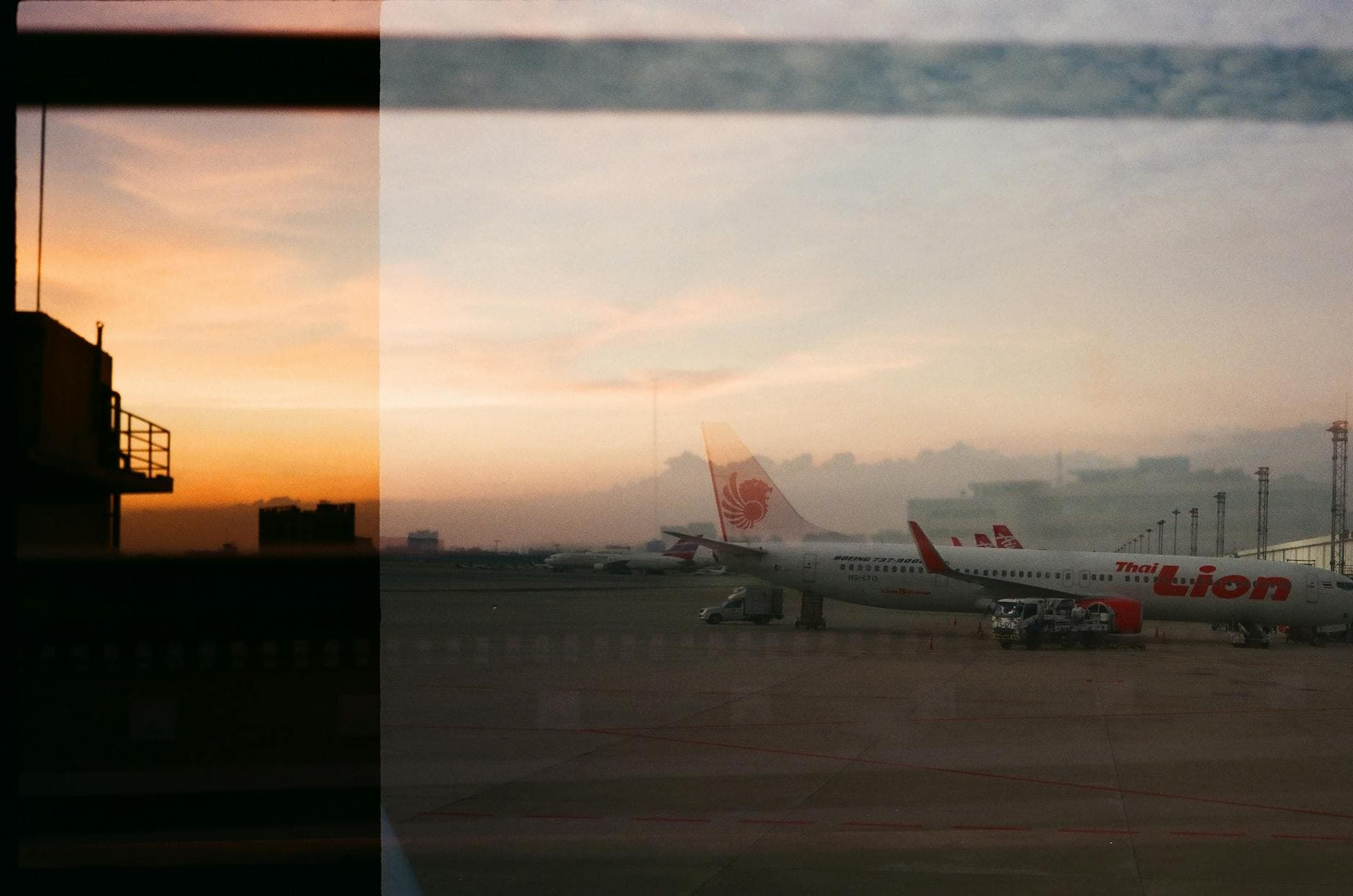 Silhouetted airplanes are lined up at the airport during sunset, seen through a window, creating a dramatic sky view.