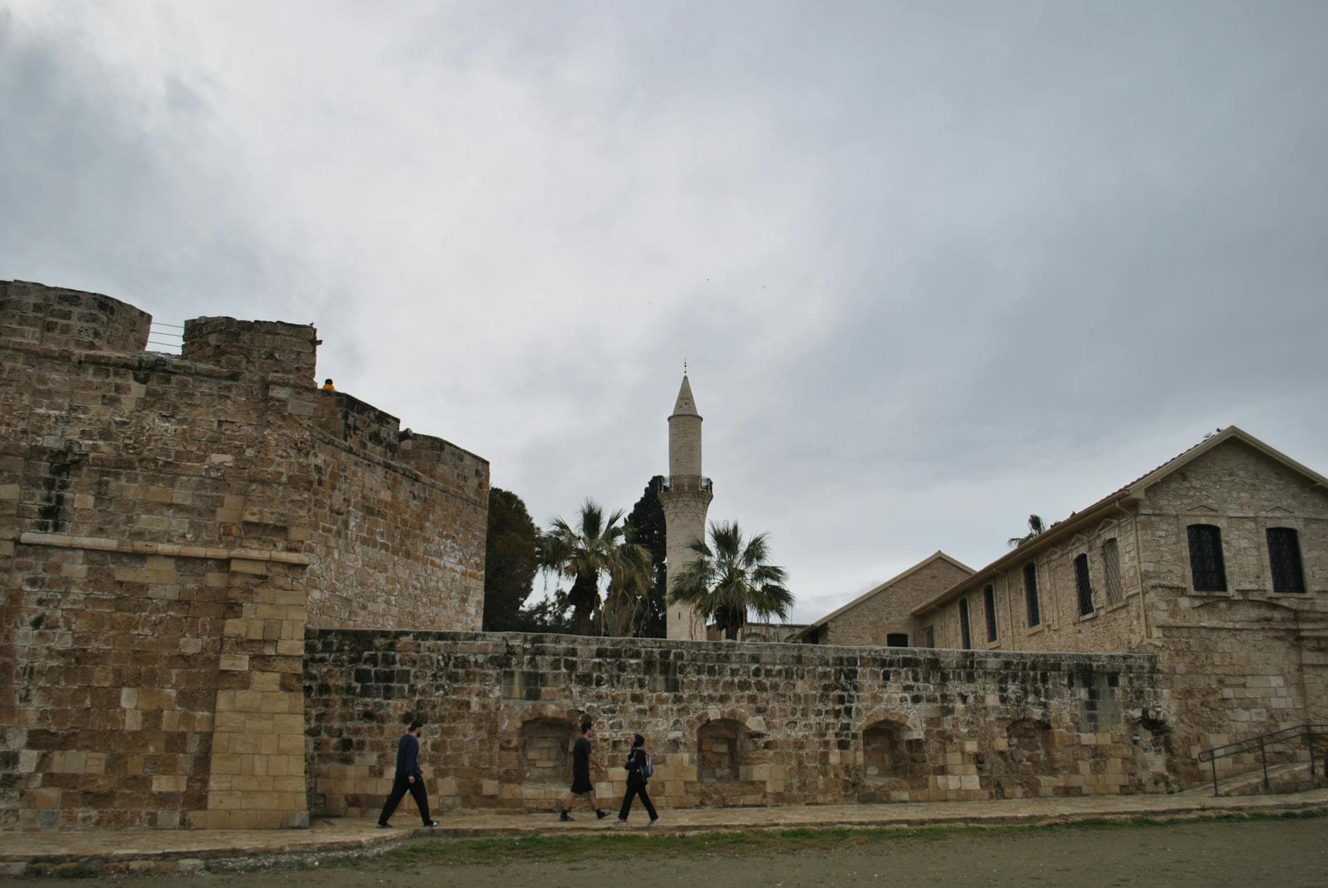 Ancient stone fortress with a prominent minaret surrounded by palm trees against a cloudy sky.