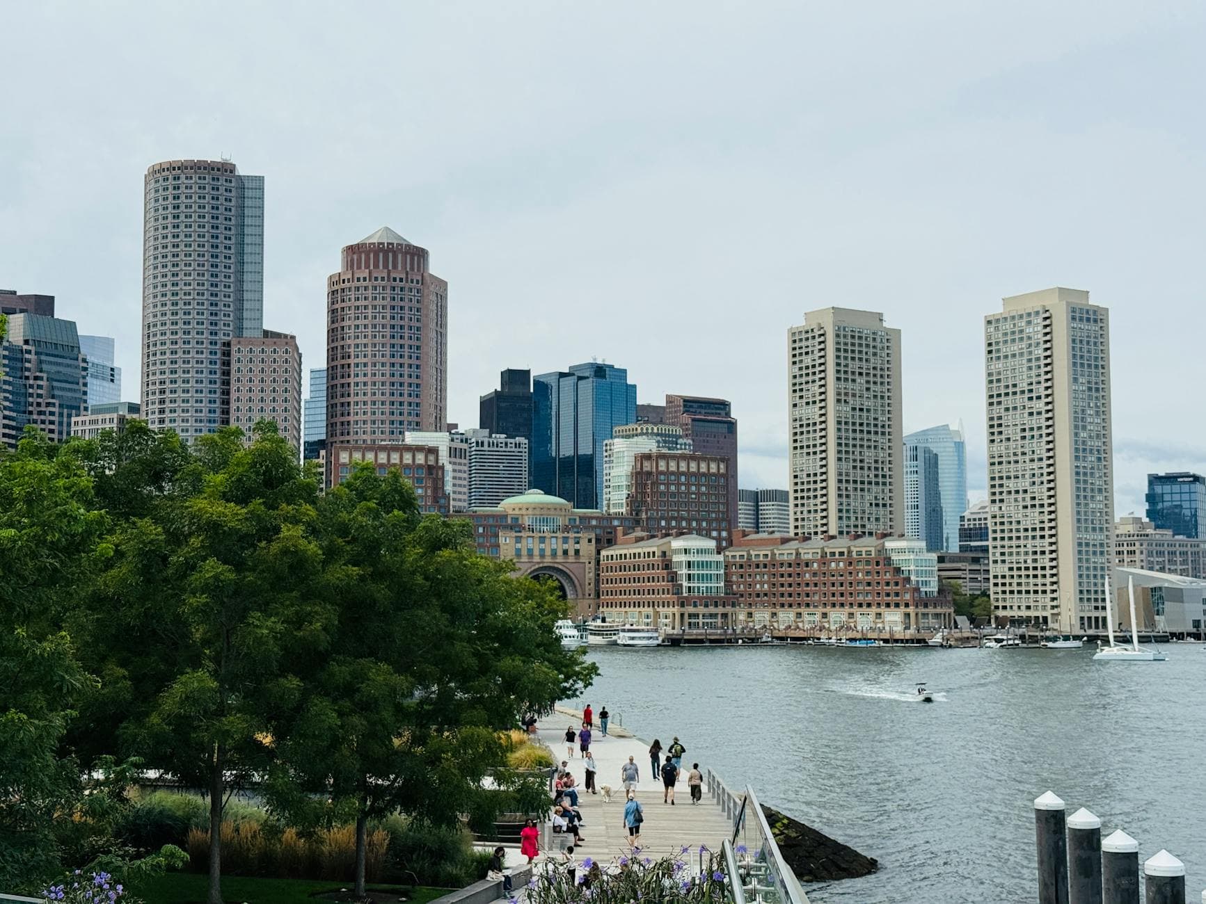 A scenic view of Boston's skyline with a bustling waterfront promenade and trees.