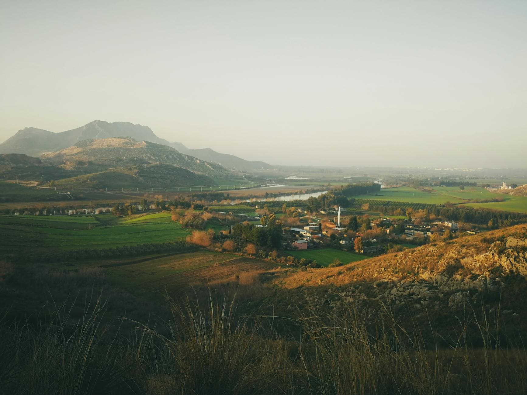 A picturesque view of Ceyhan in Adana, Turkey, featuring vast fields, a village, and distant mountains under a morning sky.