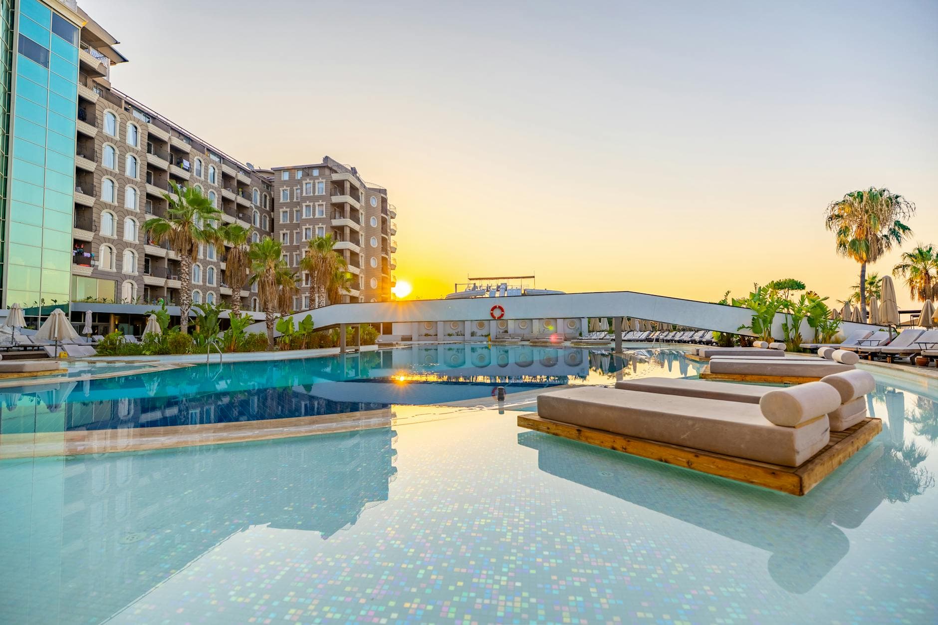 Stunning sunset view of a luxury resort pool in Didim, Türkiye, with palm trees and modern architecture.