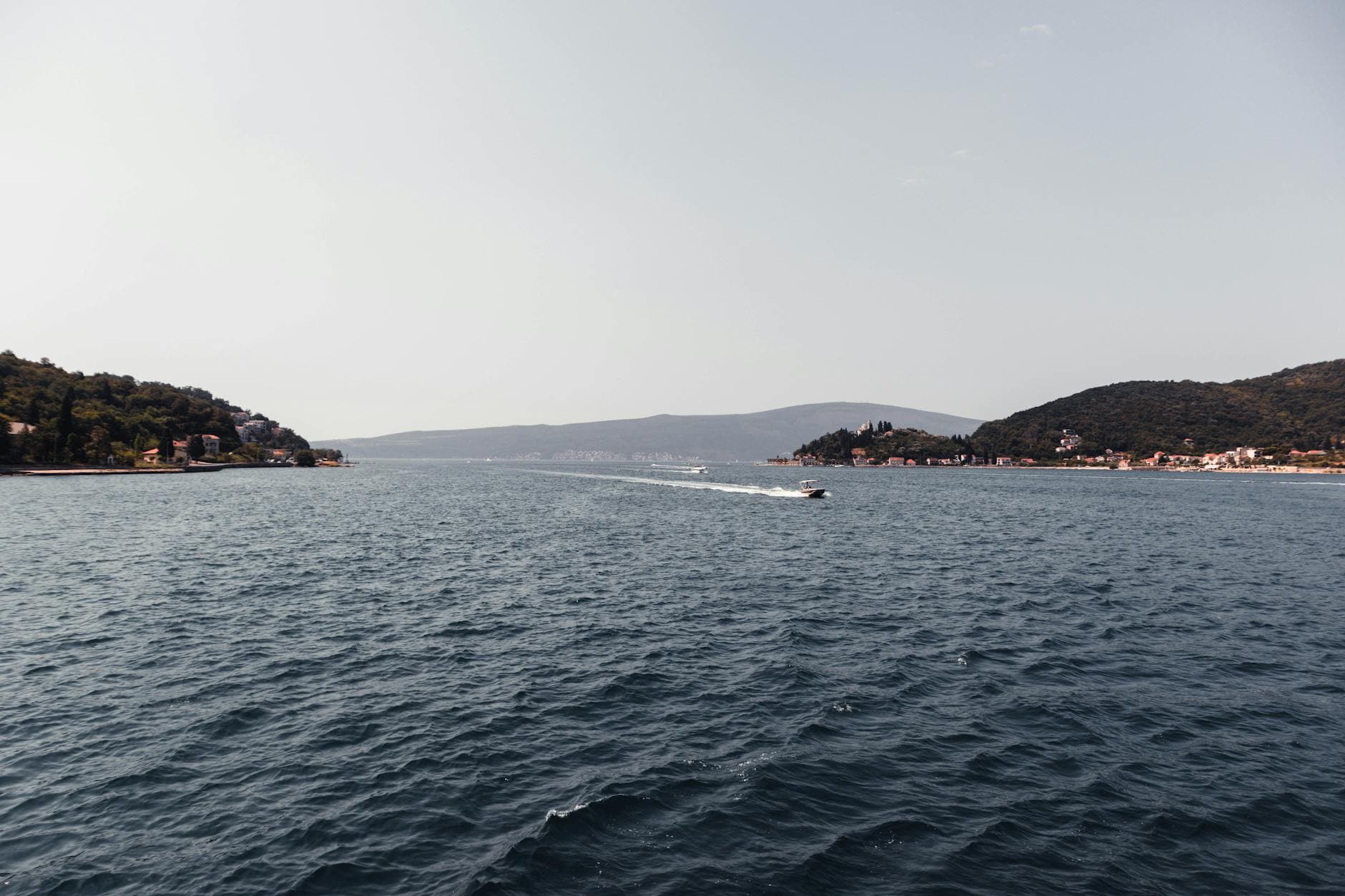 A serene view of the Herceg Novi Bay with a boat cutting through the tranquil waters, surrounded by lush hills.