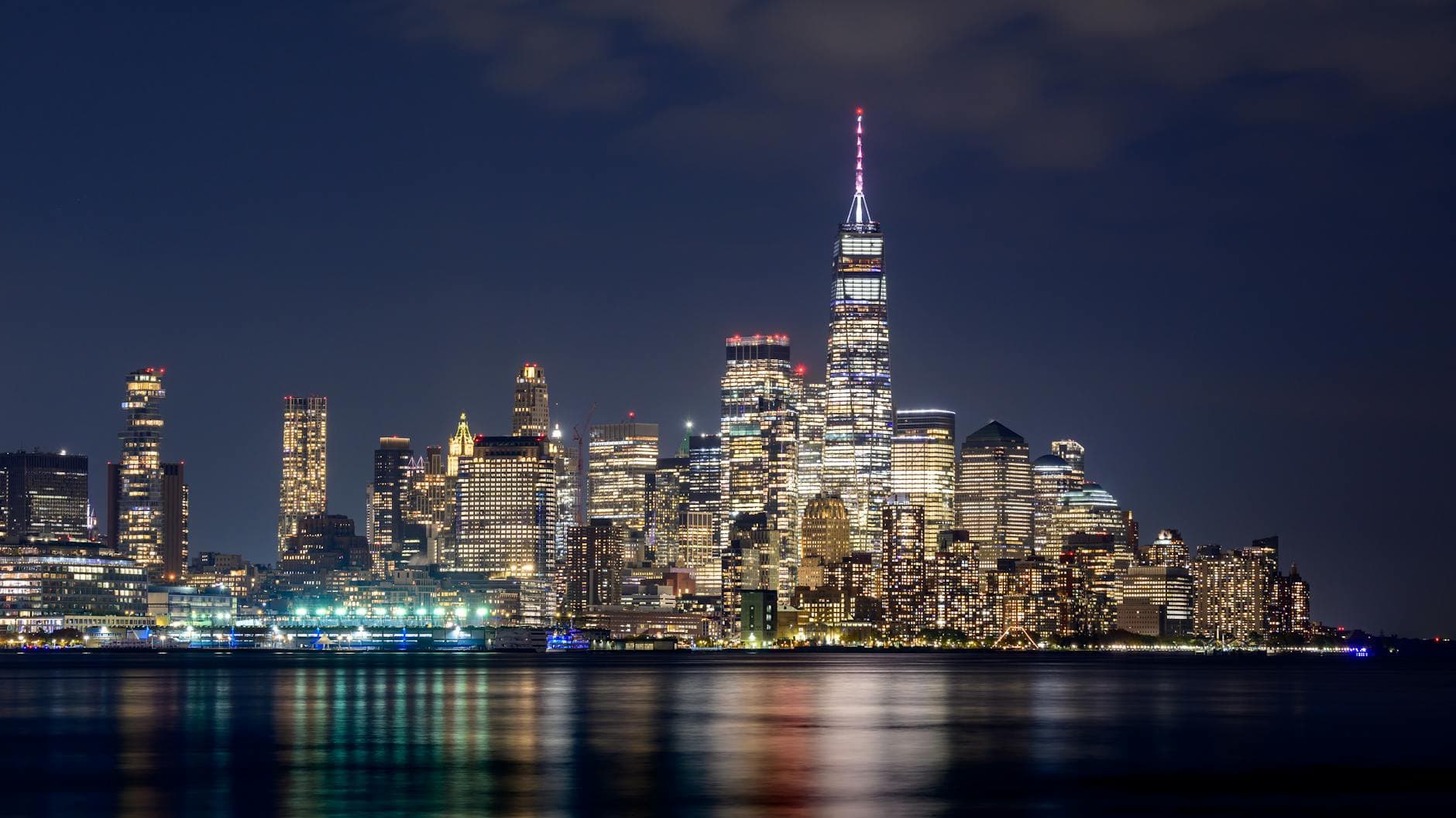 Dazzling New York City skyline at night with illuminated skyscrapers, featuring the iconic One World Trade Center reflecting in the water.