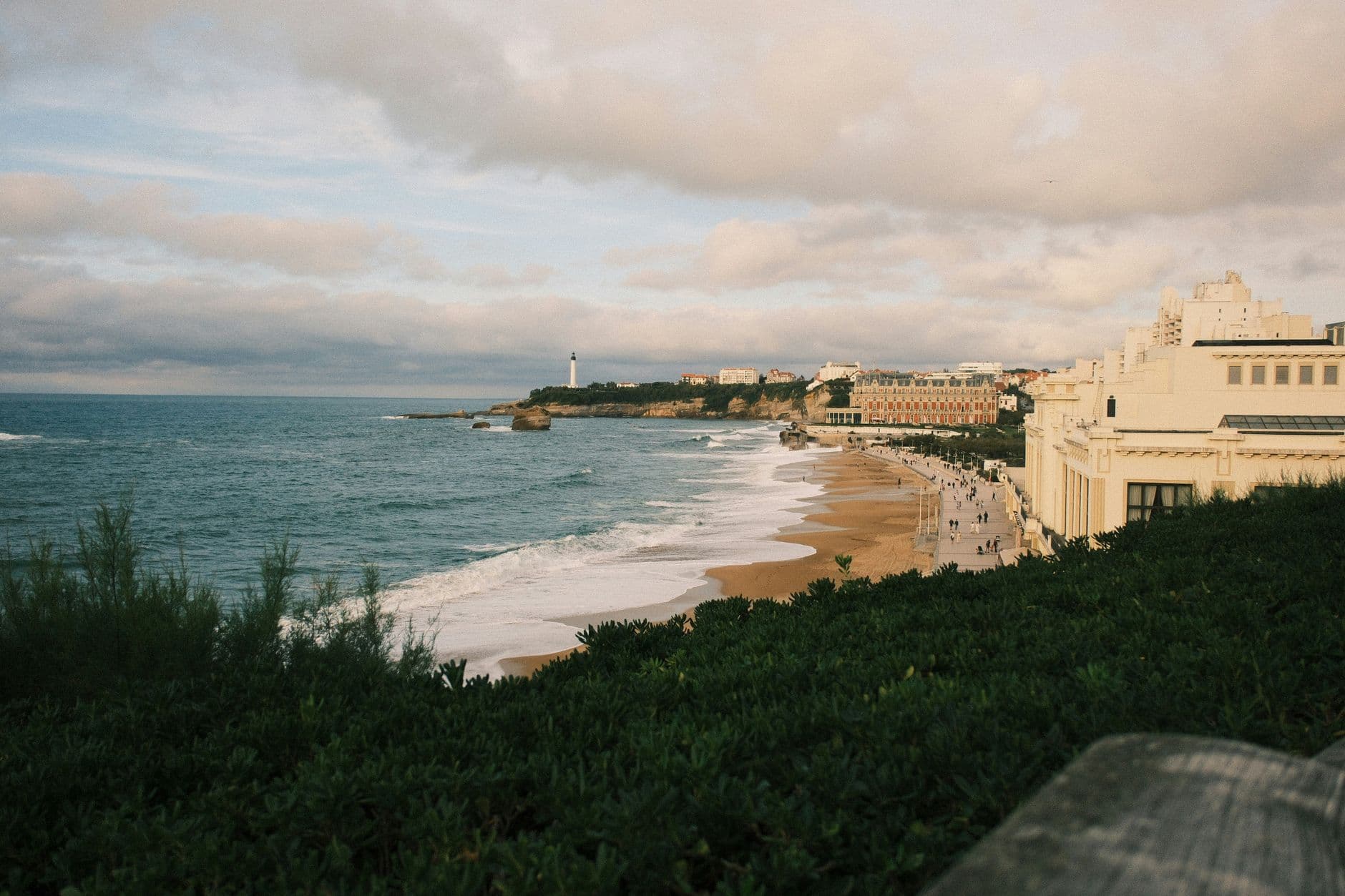 Explore Biarritz's scenic coastal view with sandy beaches and lighthouse under a cloudy sky.