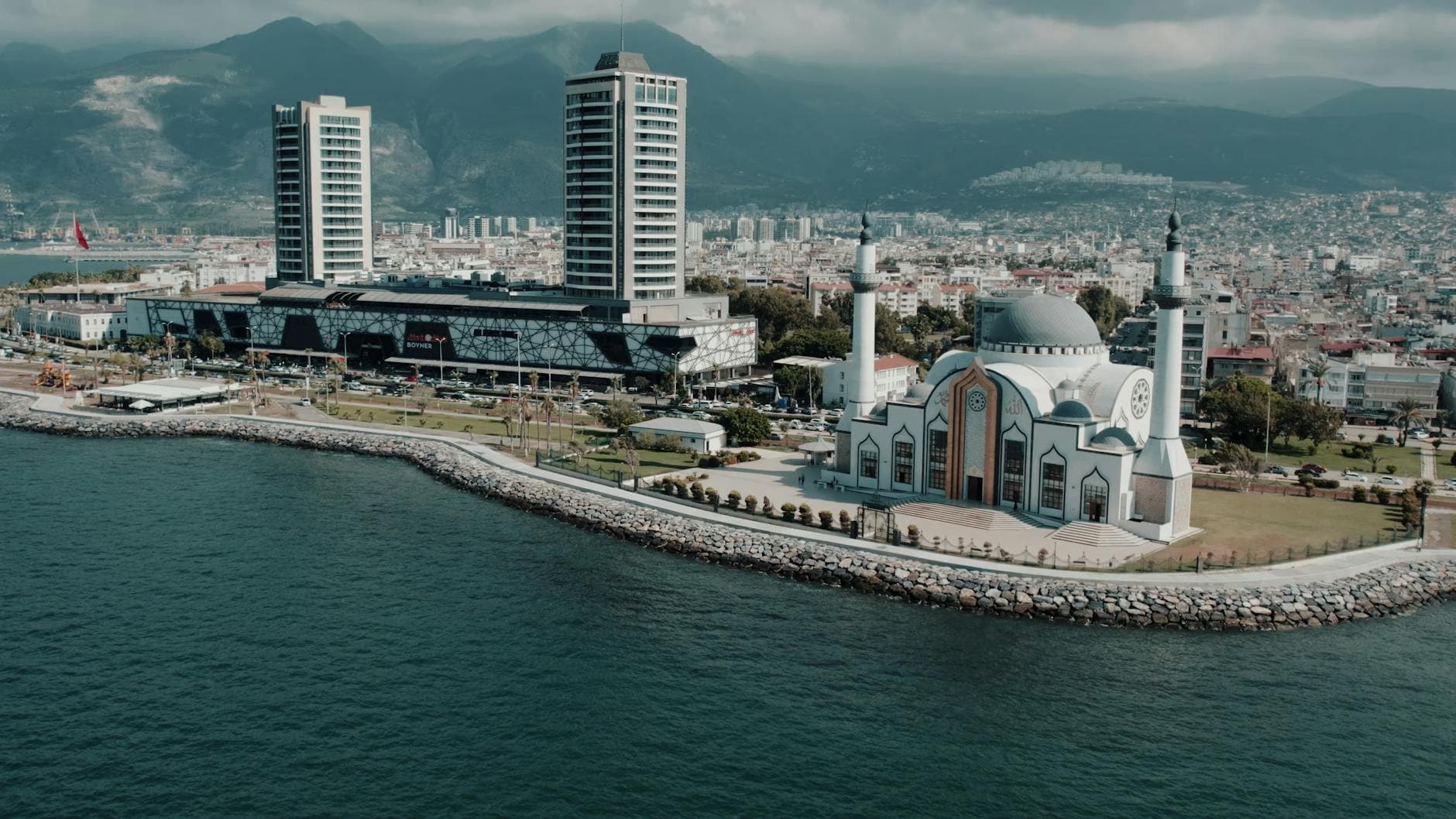 Aerial photograph showcasing İskenderun's mosque and cityscape against the mountainous backdrop on a sunny day.
