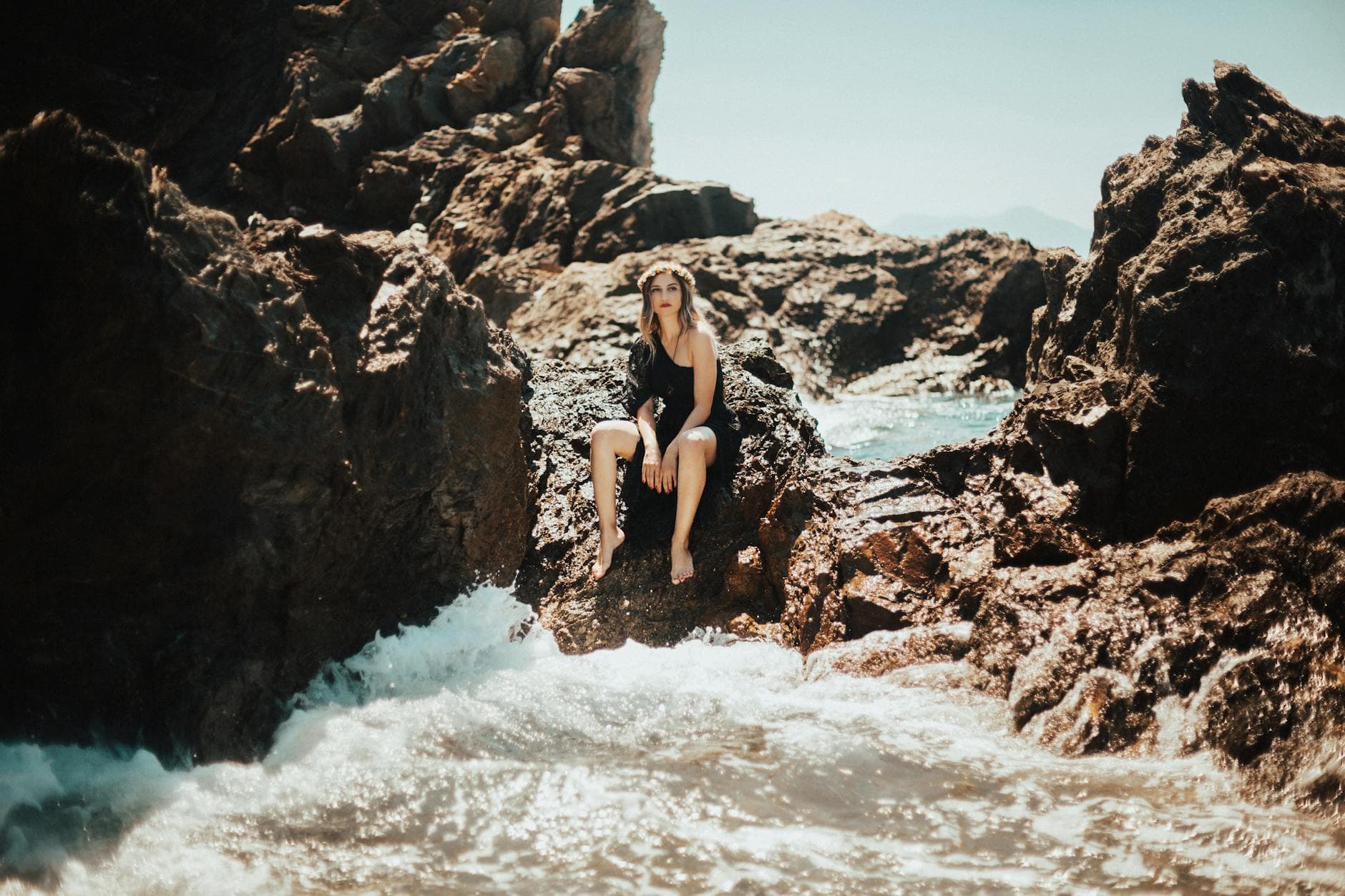 A woman in a black dress sitting on rocky shore with ocean waves in Karaburun, Turkey.