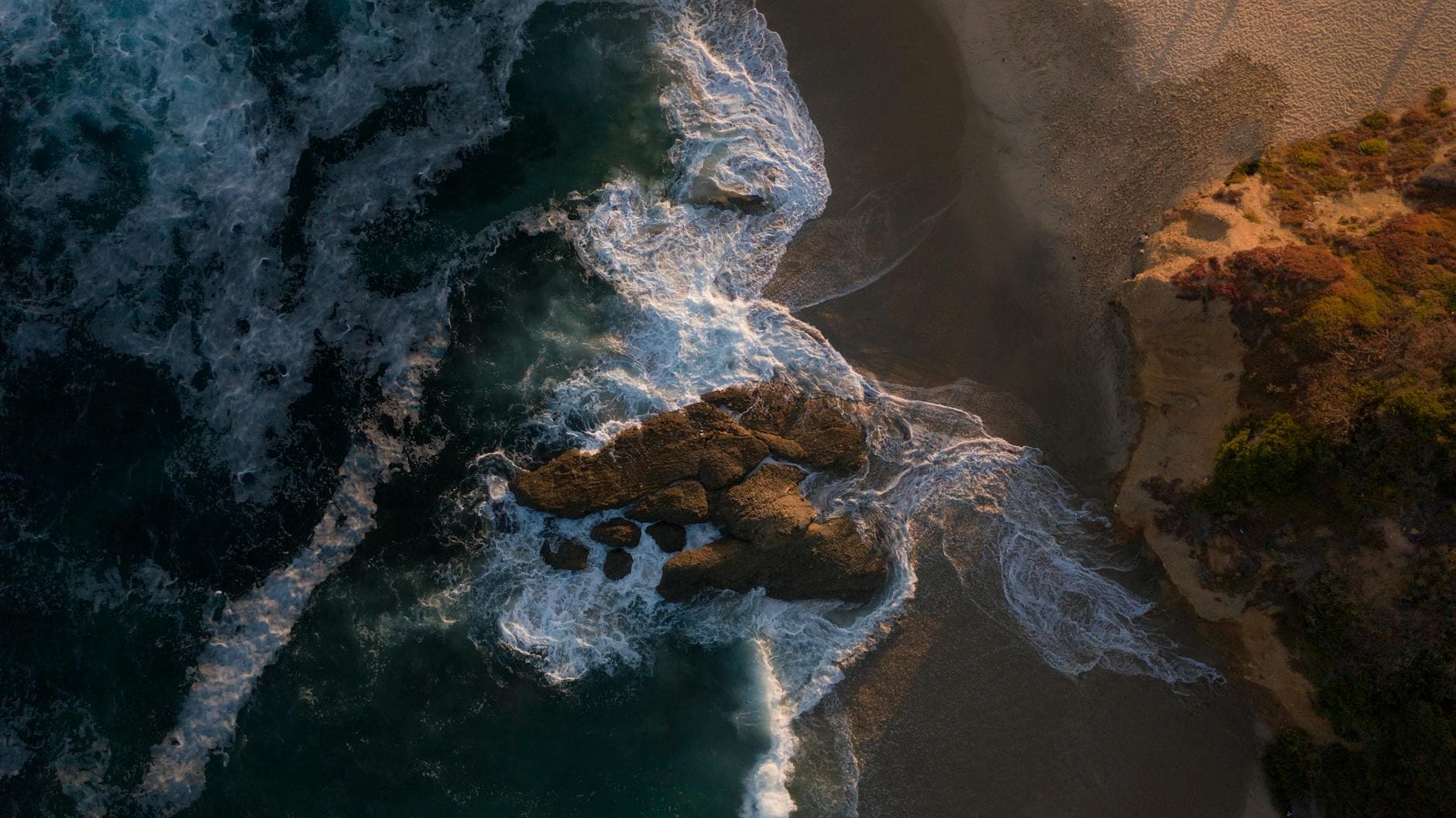 Aerial shot of Laguna Beach coastline with waves crashing on rocks and white sand.