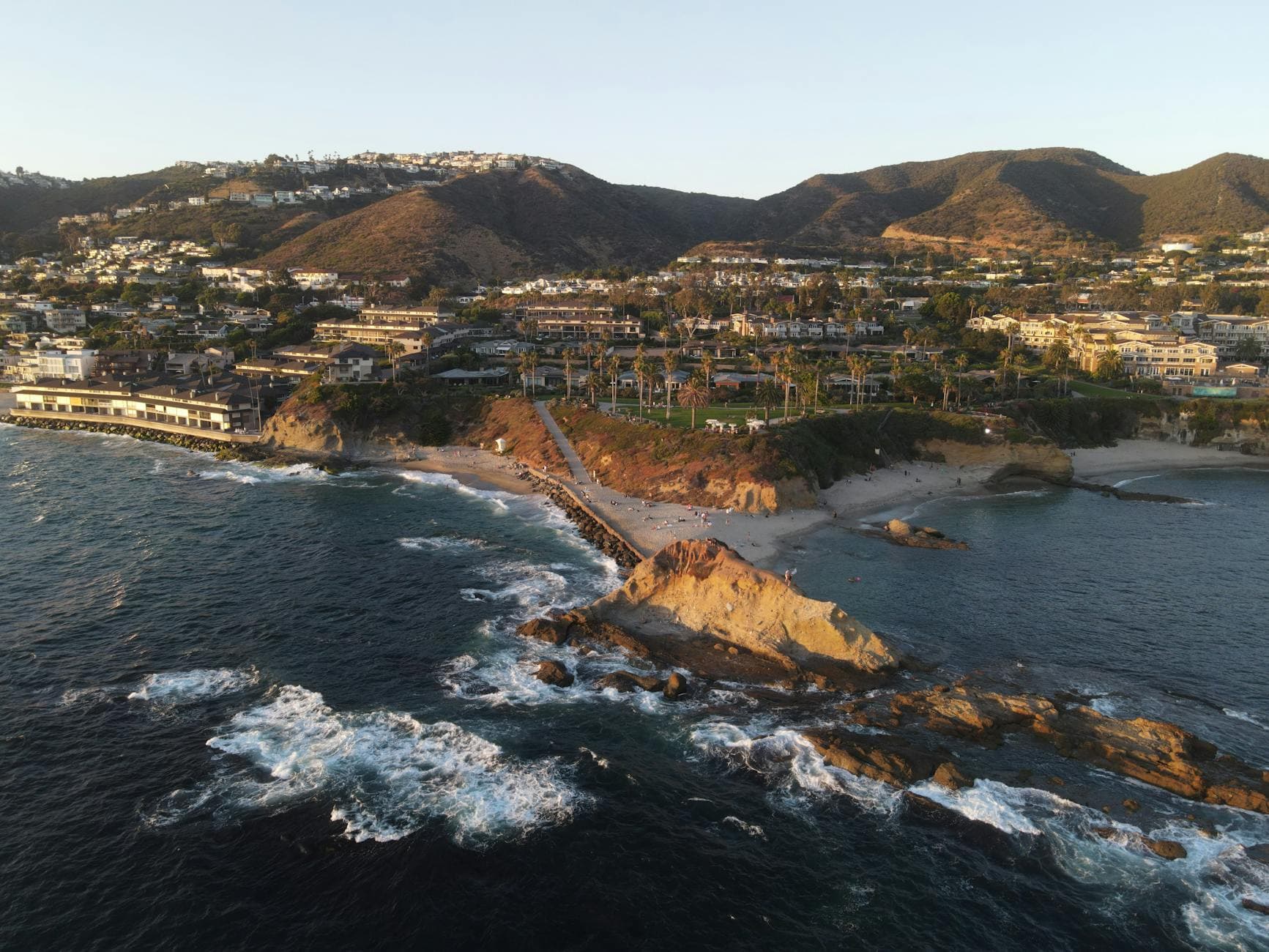 Stunning aerial view of Laguna Beach's coastline with houses, rocky shore, and ocean waves during sunset.