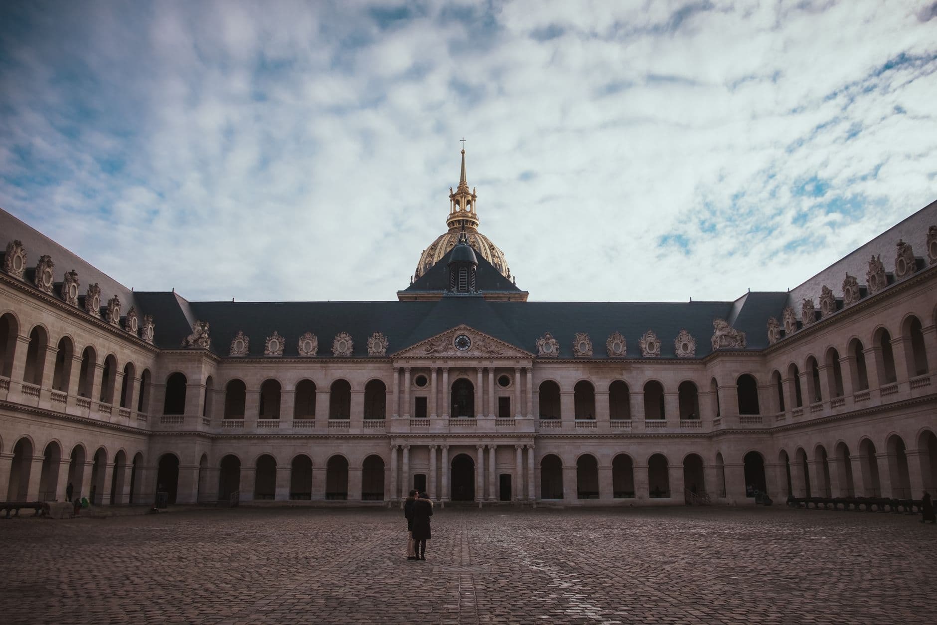 Historic courtyard of Les Invalides with a view of the chapel dome under a cloudy winter sky.