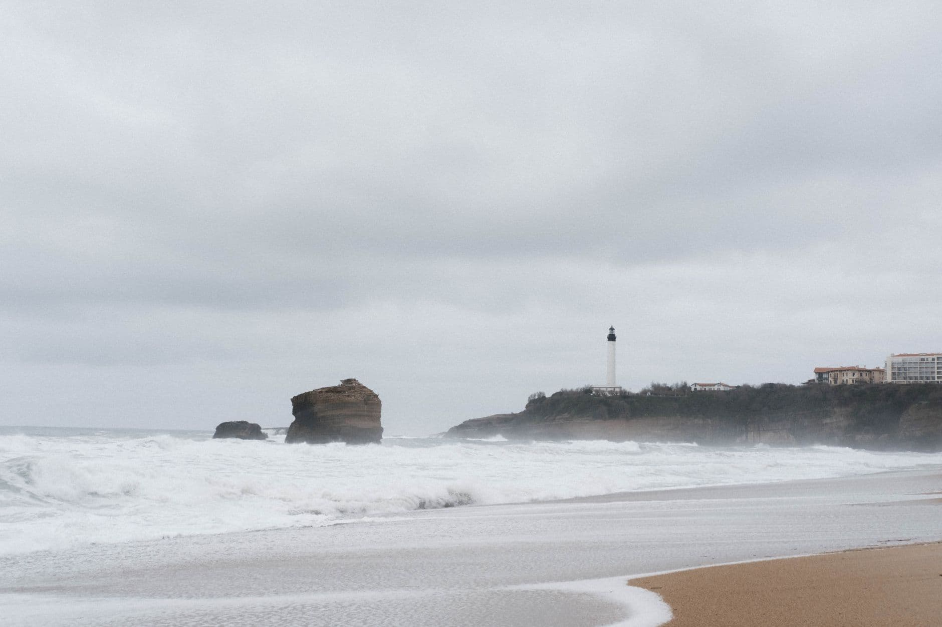 Scenic view of Biarritz beach with a lighthouse and ocean waves under a cloudy sky.
