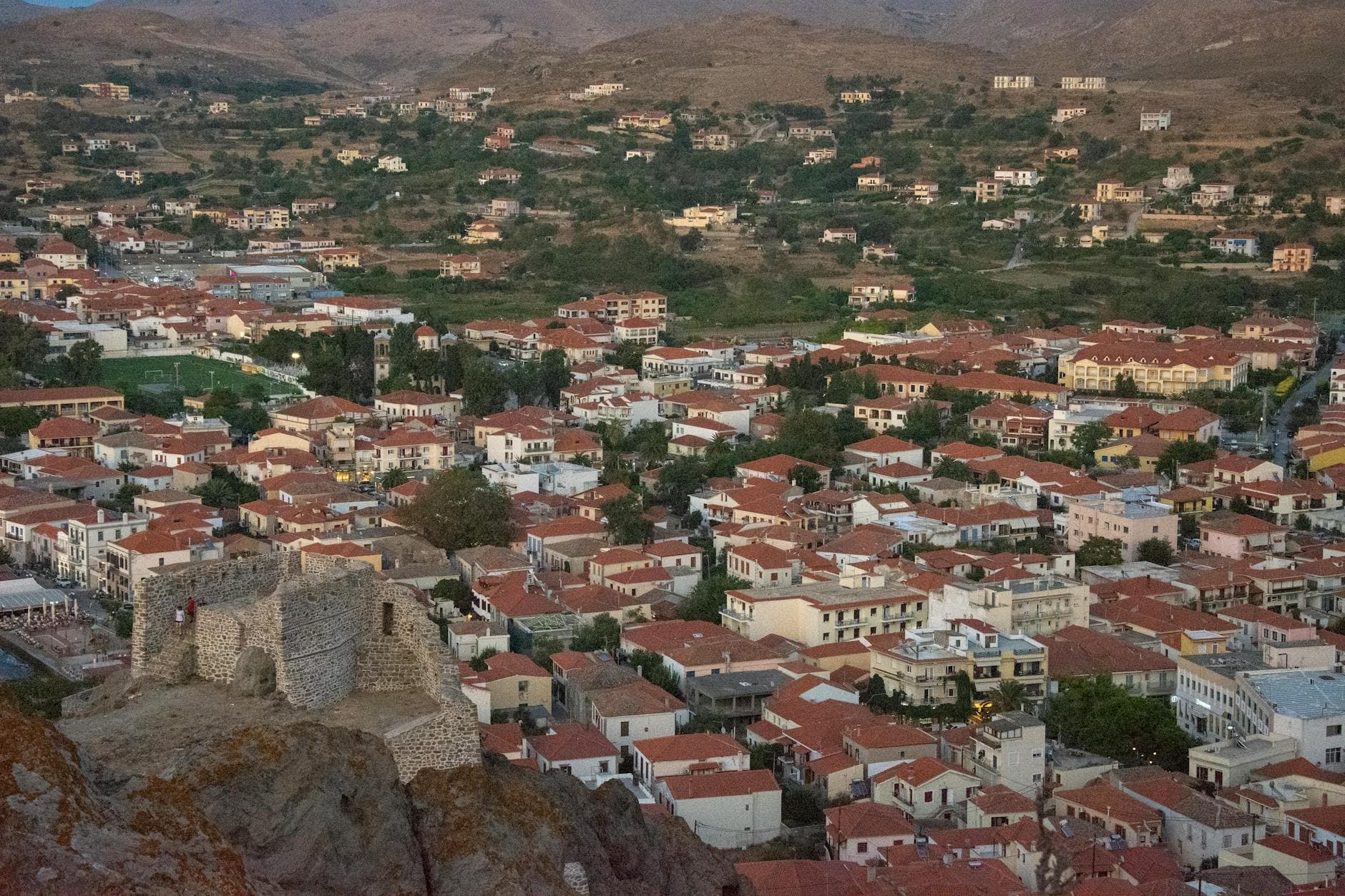 Aerial view showcasing a historic town with red rooftops and ancient castle ruins in a mountainous landscape.