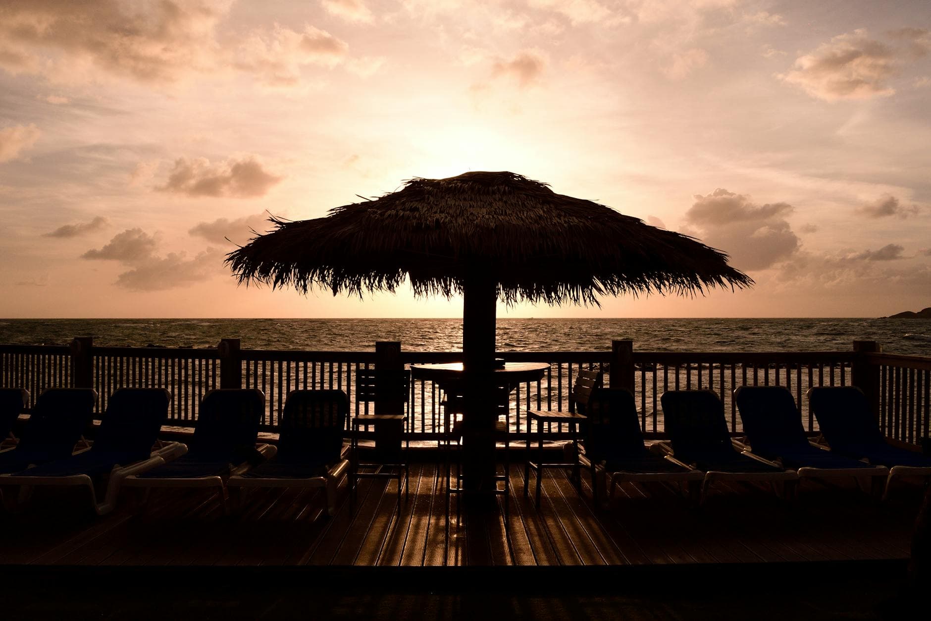 Silhouette of a thatched roof umbrella and loungers at sunset on a St. Lucia beach deck.