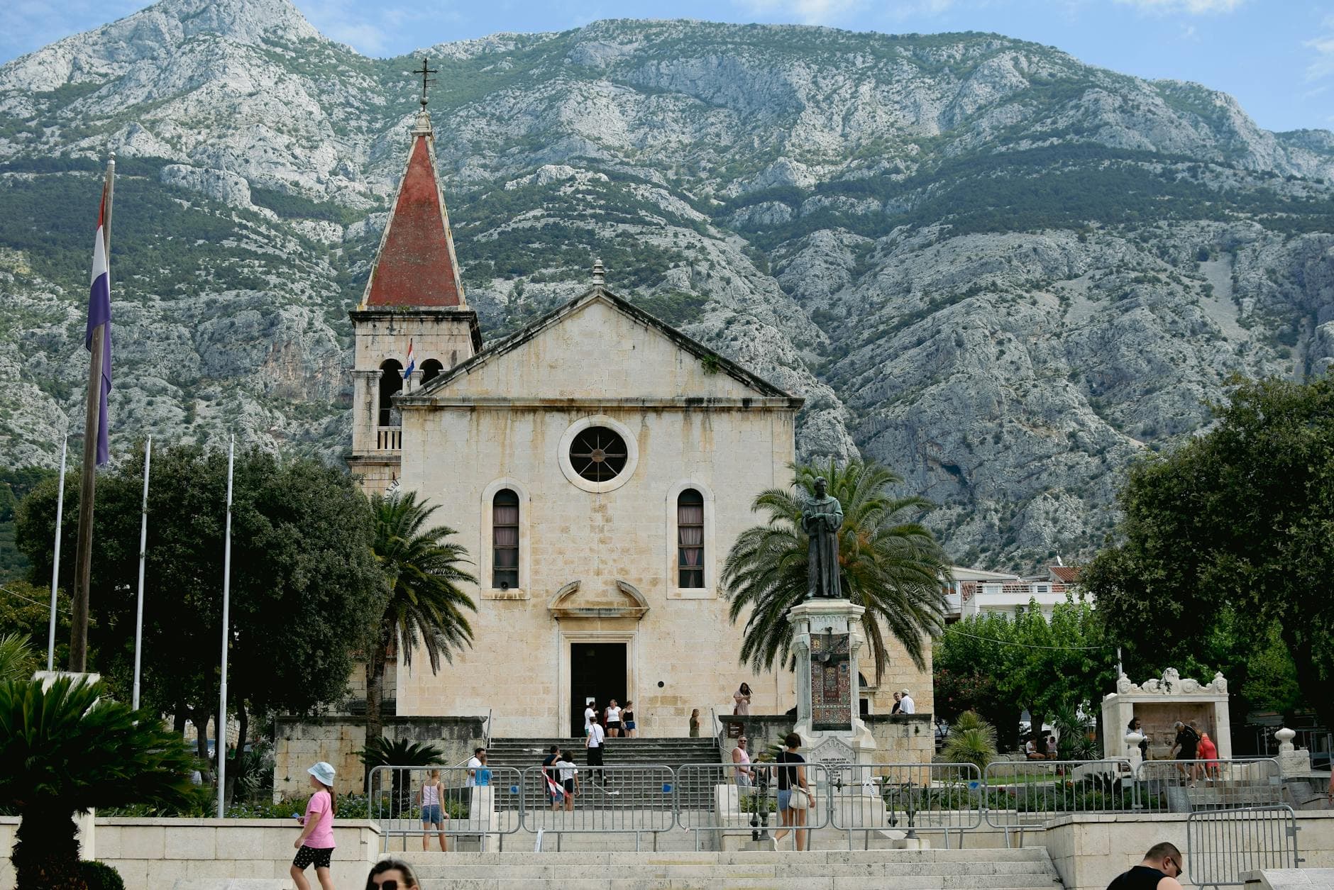 A scenic view of a historic church in Makarska, Croatia, surrounded by mountains and summer tourists.