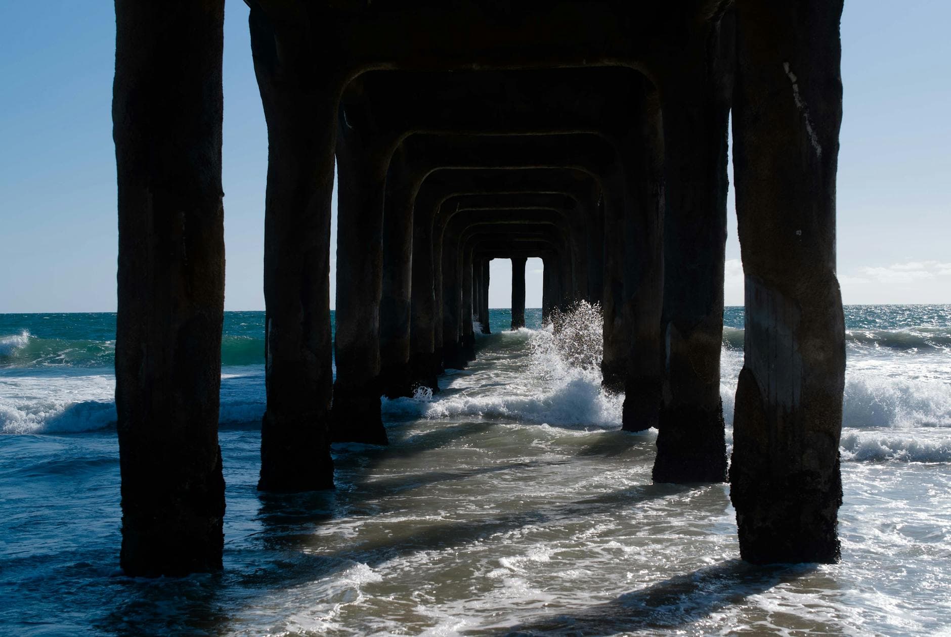 View under Manhattan Beach Pier as waves crash against the structure, capturing the ocean's power.