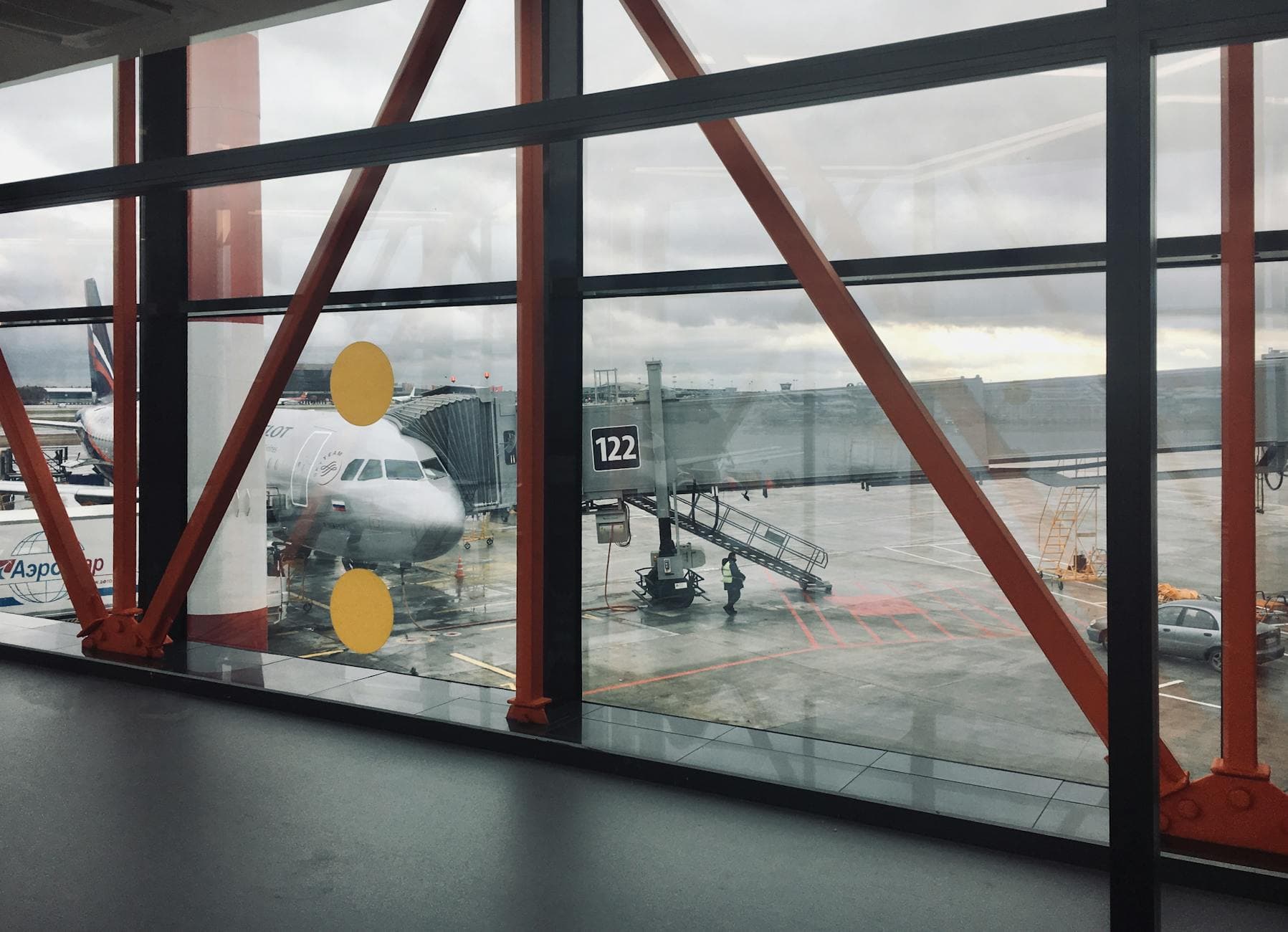 View of a commercial airplane parked at an airport terminal through large glass windows, showcasing airport activity.