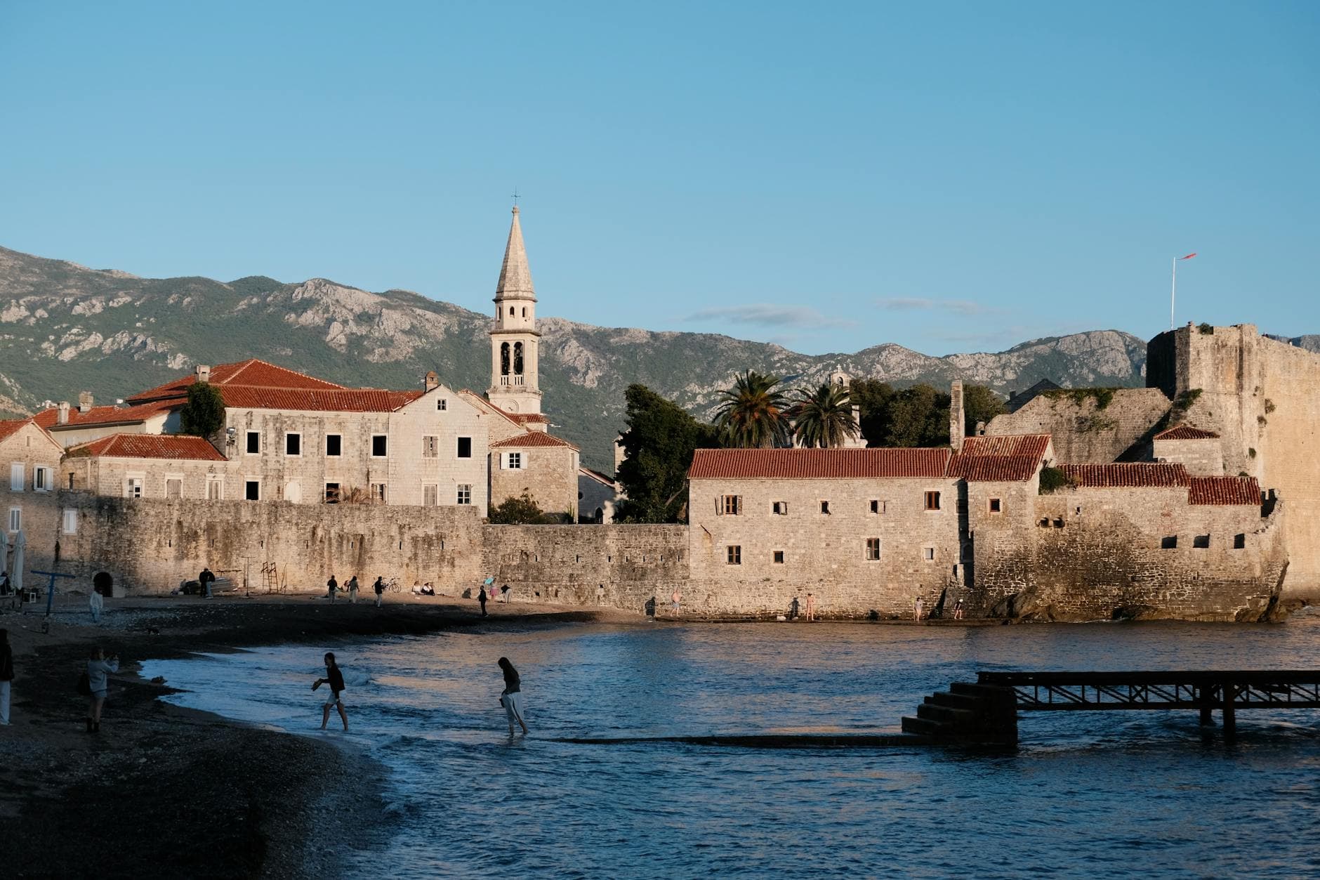 Charming seaside view of Budva's ancient walls during sunset with people enjoying the beach.