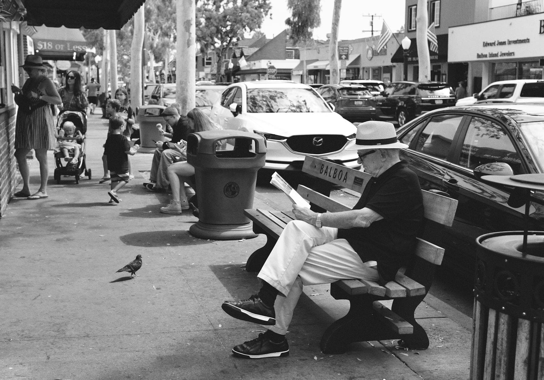 A lively street scene in Balboa, Newport Beach, featuring people interacting and a senior adult reading on a bench.