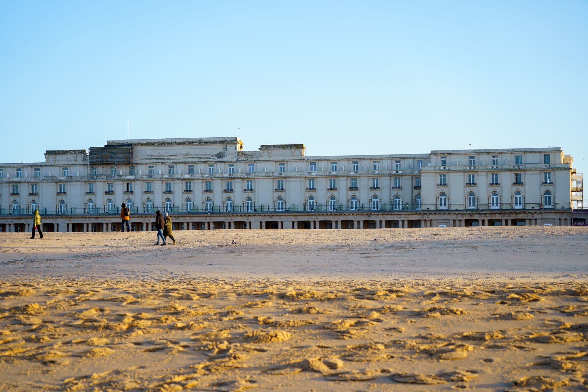 Historic building facade seen from a sandy beach during a sunny day with people strolling.