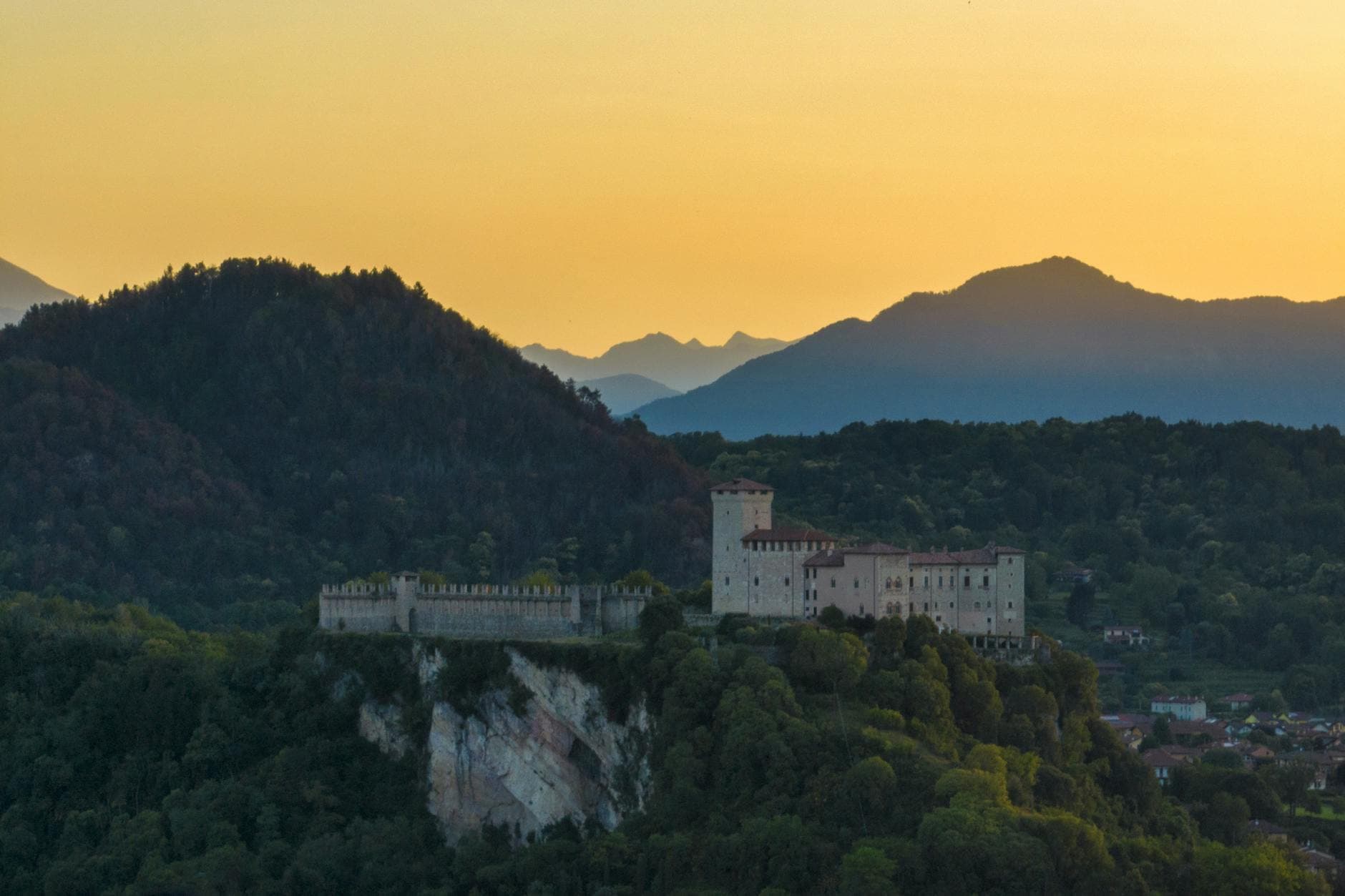 A scenic view of a historic Italian castle on a hill at sunset, surrounded by lush green hills.