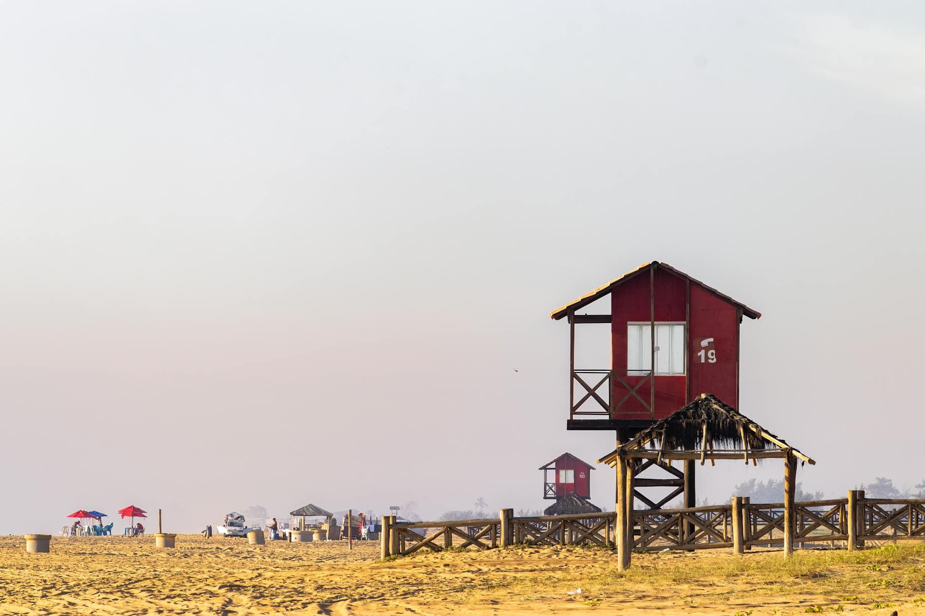 Red lifeguard tower on a sunny Brazilian beach with white sky and umbrellas in view.