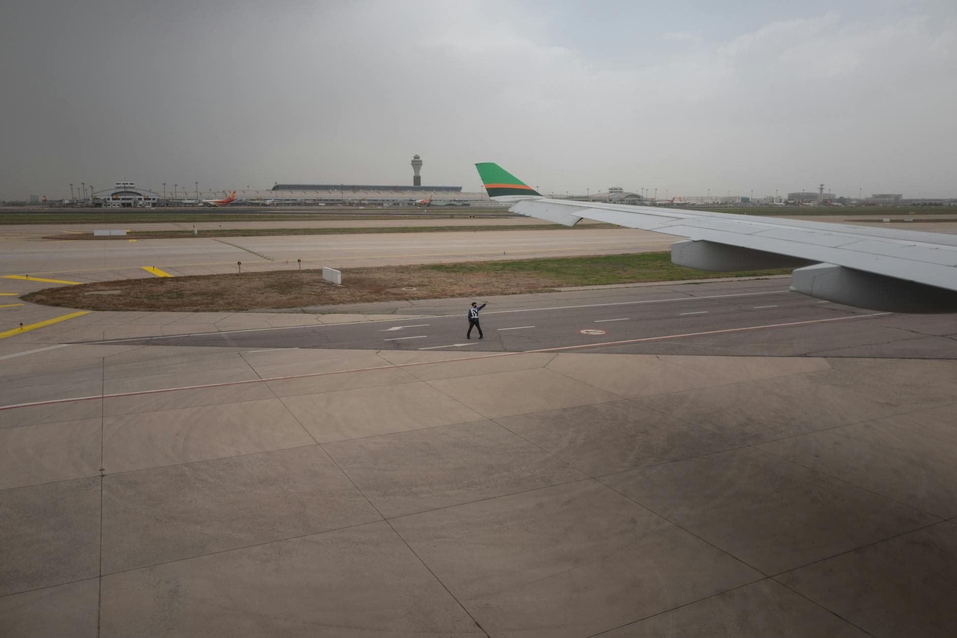 View of an airport runway with aircraft wing, control tower, and ground staff guiding on a cloudy day.