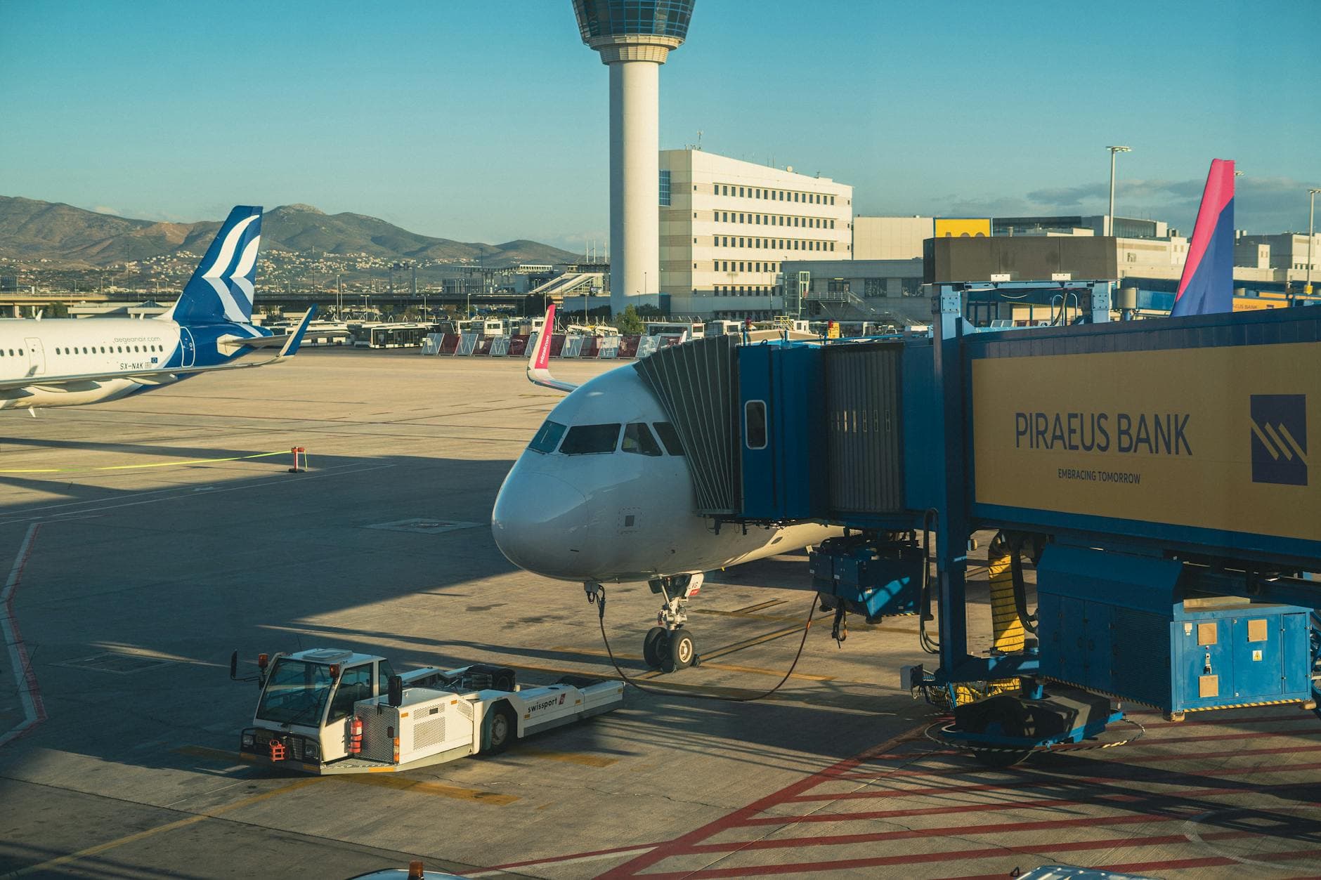 An airplane at Athens International Airport gate during a sunny day, featuring a jet bridge and terminal view.