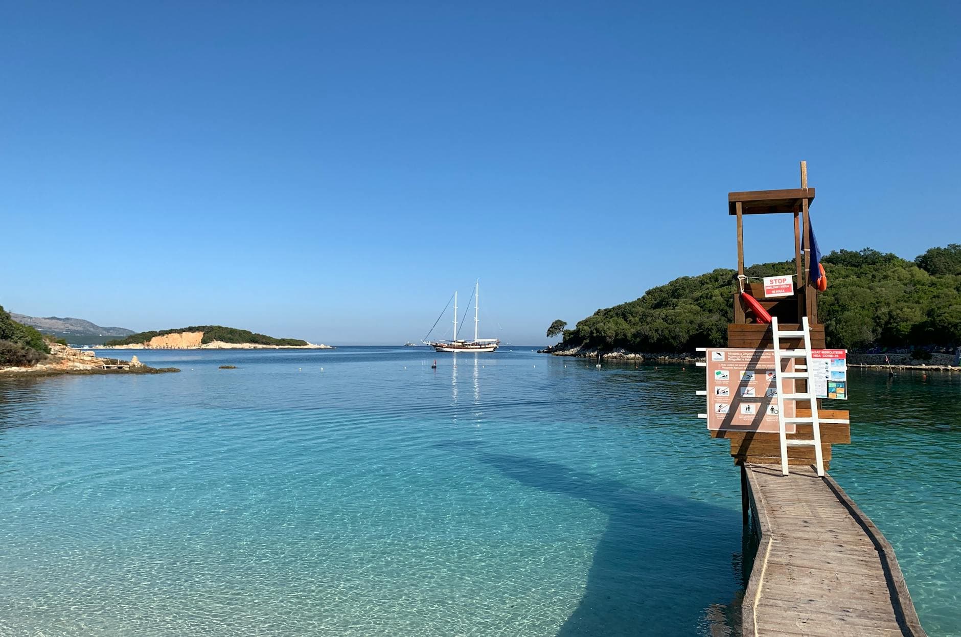 Idyllic view of Albanian coastline featuring a sailboat and lifeguard tower under clear skies. Perfect summer day.