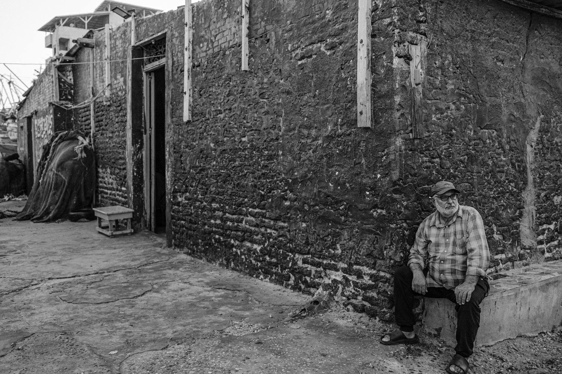 Black and white portrait of an elderly man sitting by a textured wall in a quaint Egyptian street.