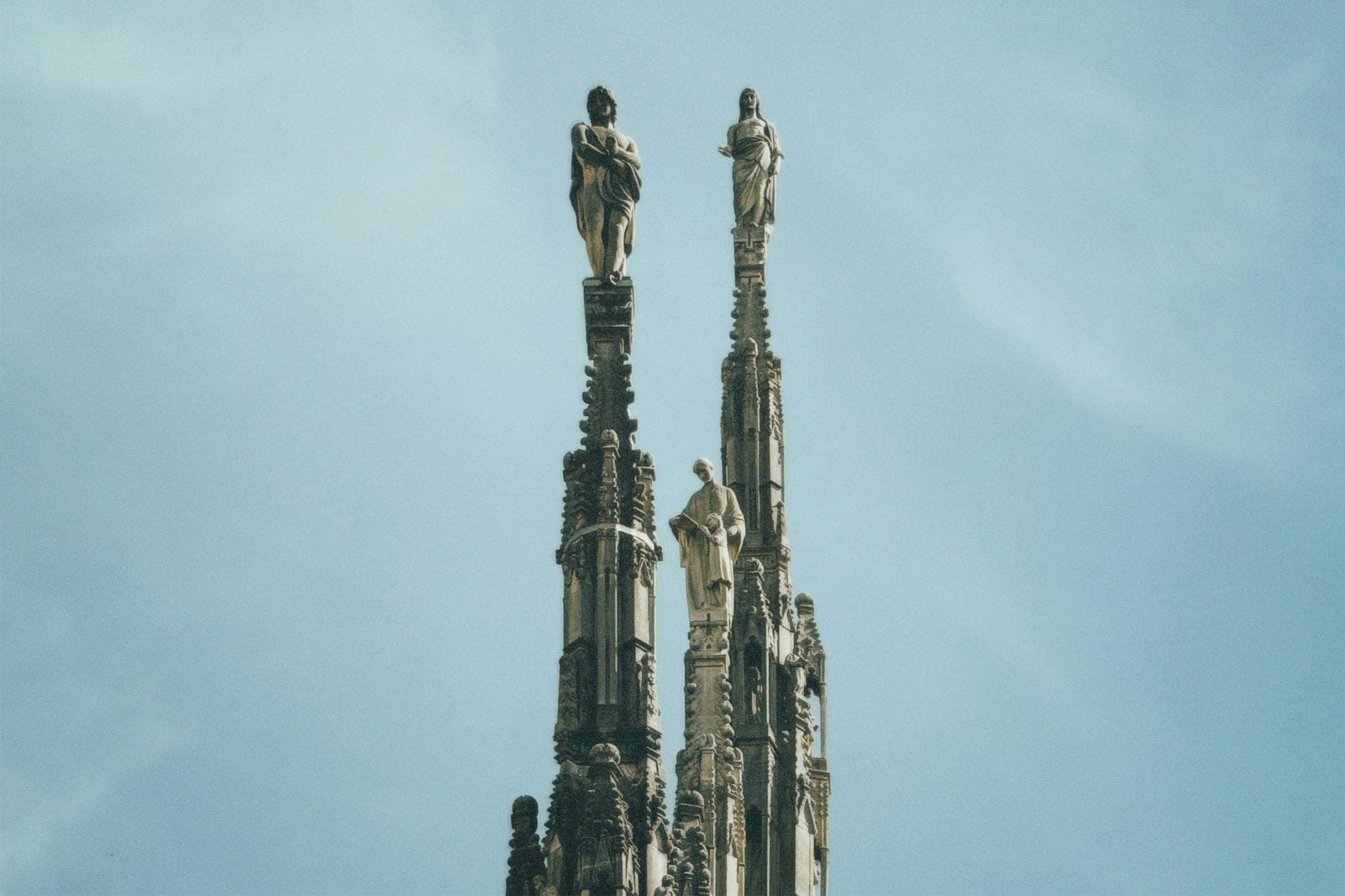 Detailed view of Gothic sculptures atop spires of the Milan Cathedral against a bright blue sky.