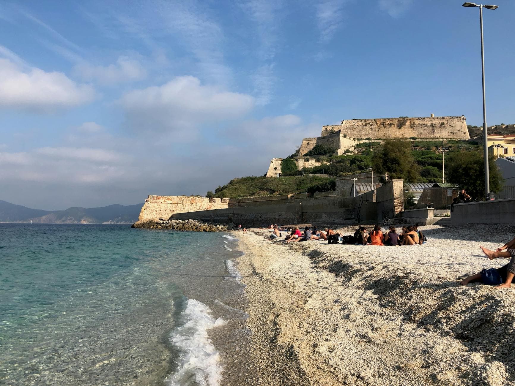 Beachgoers enjoy a sunny day at Forte Stella, Portoferraio, with tranquil sea and historic architecture.