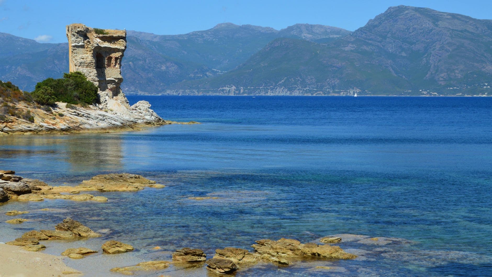 Scenic coastline in Corsica featuring historical tower ruins and mountainous backdrop.