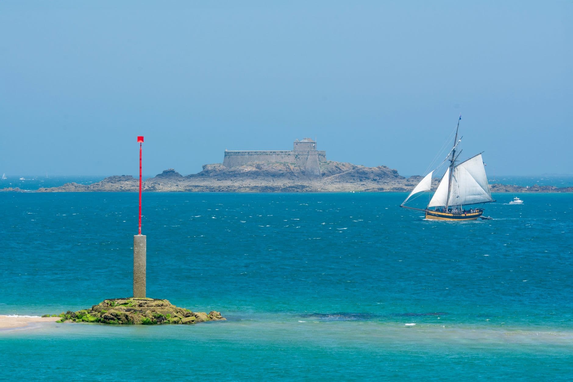 Explore the scenic Saint-Malo coast with a majestic sailing ship and historic fort in view.