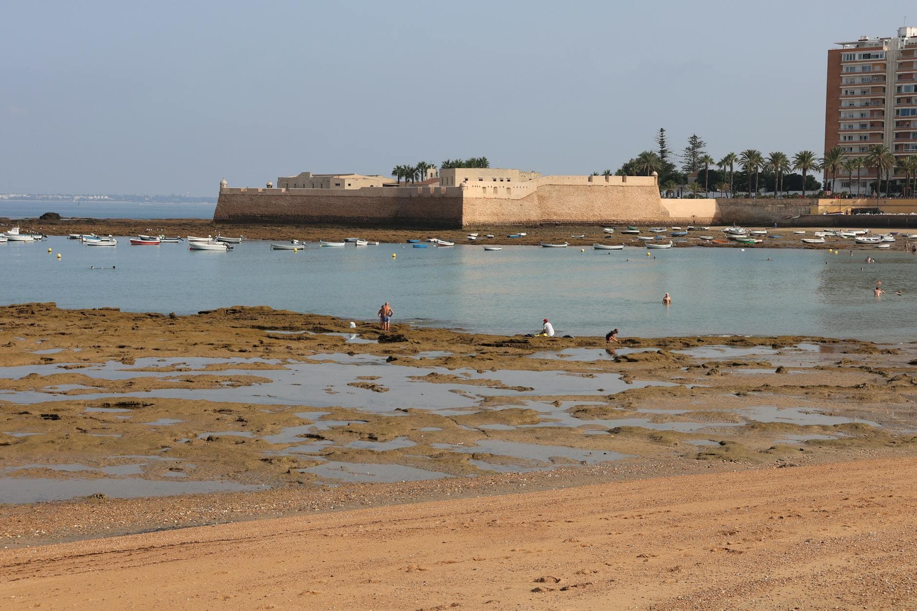 Scenic view of a historic fortress by the sea in Cádiz, featuring a serene beach and boats.