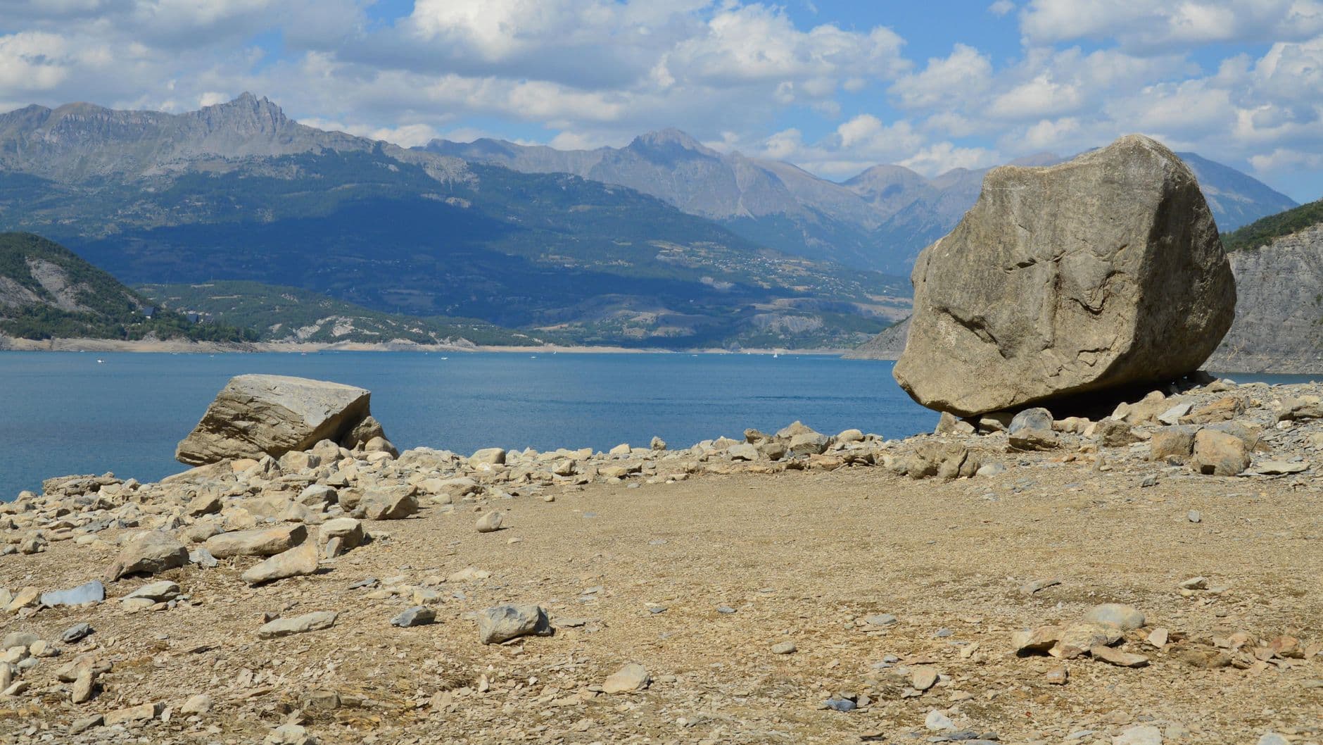Tranquil mountain lake view with large boulders on a rocky shore under a blue sky.