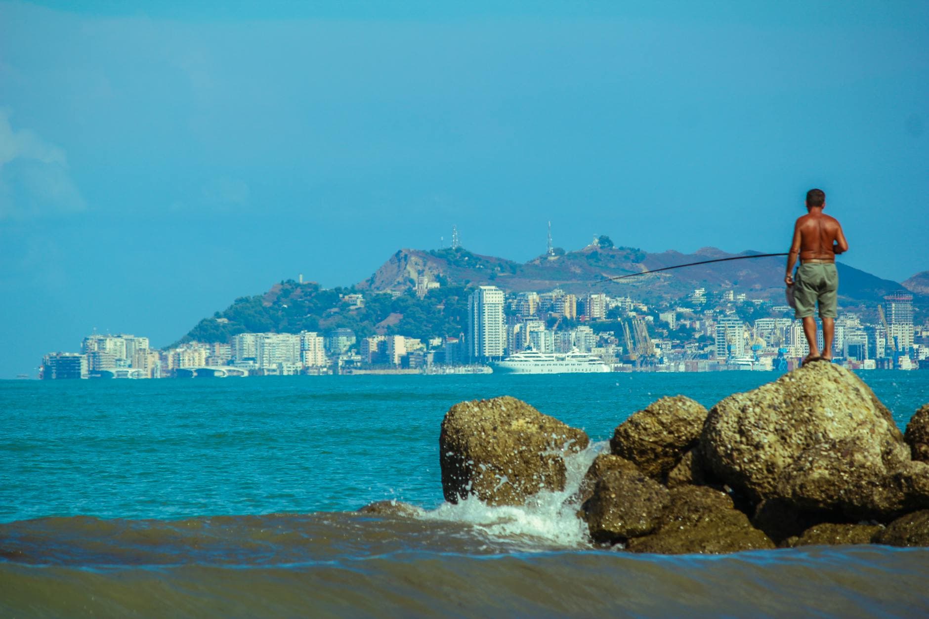 A man fishes on coastal rocks with Durrës cityscape in the background, Albania.