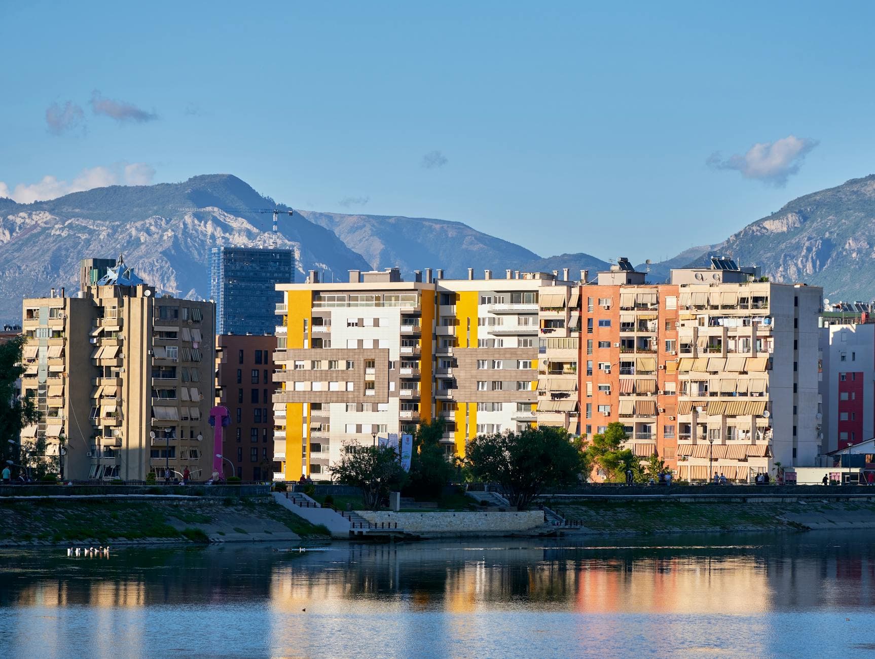 Colorful apartment buildings by a waterfront in Tirana, Albania, with mountains in the background.