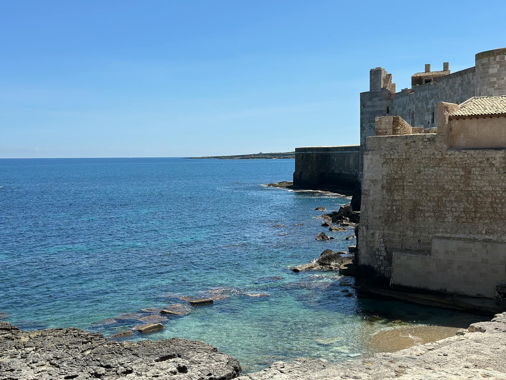 Scenic view of Ortigia's coastline in Sicily with historical stone buildings against clear blue waters.