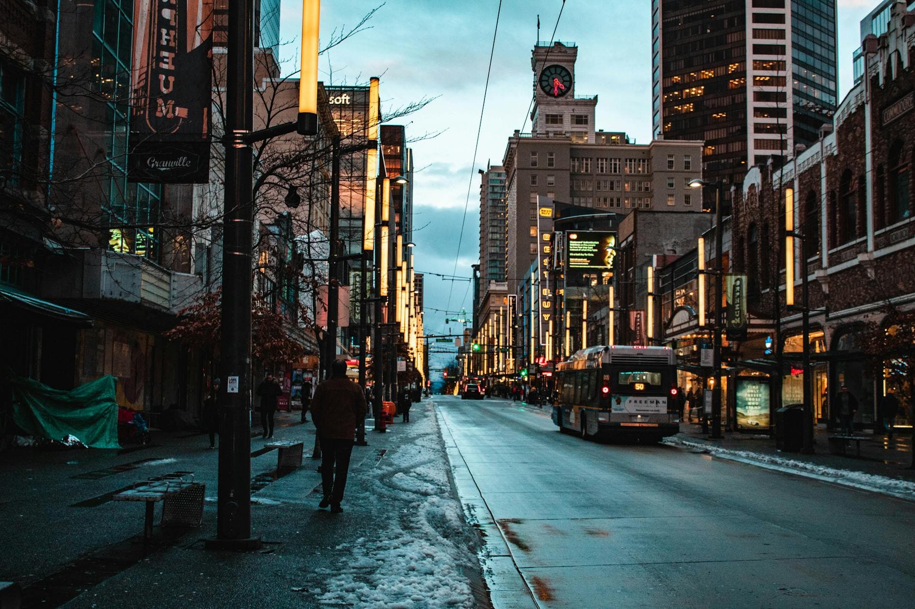 Urban city street view of Vancouver at dusk featuring a bus, clock tower, and high-rise buildings.