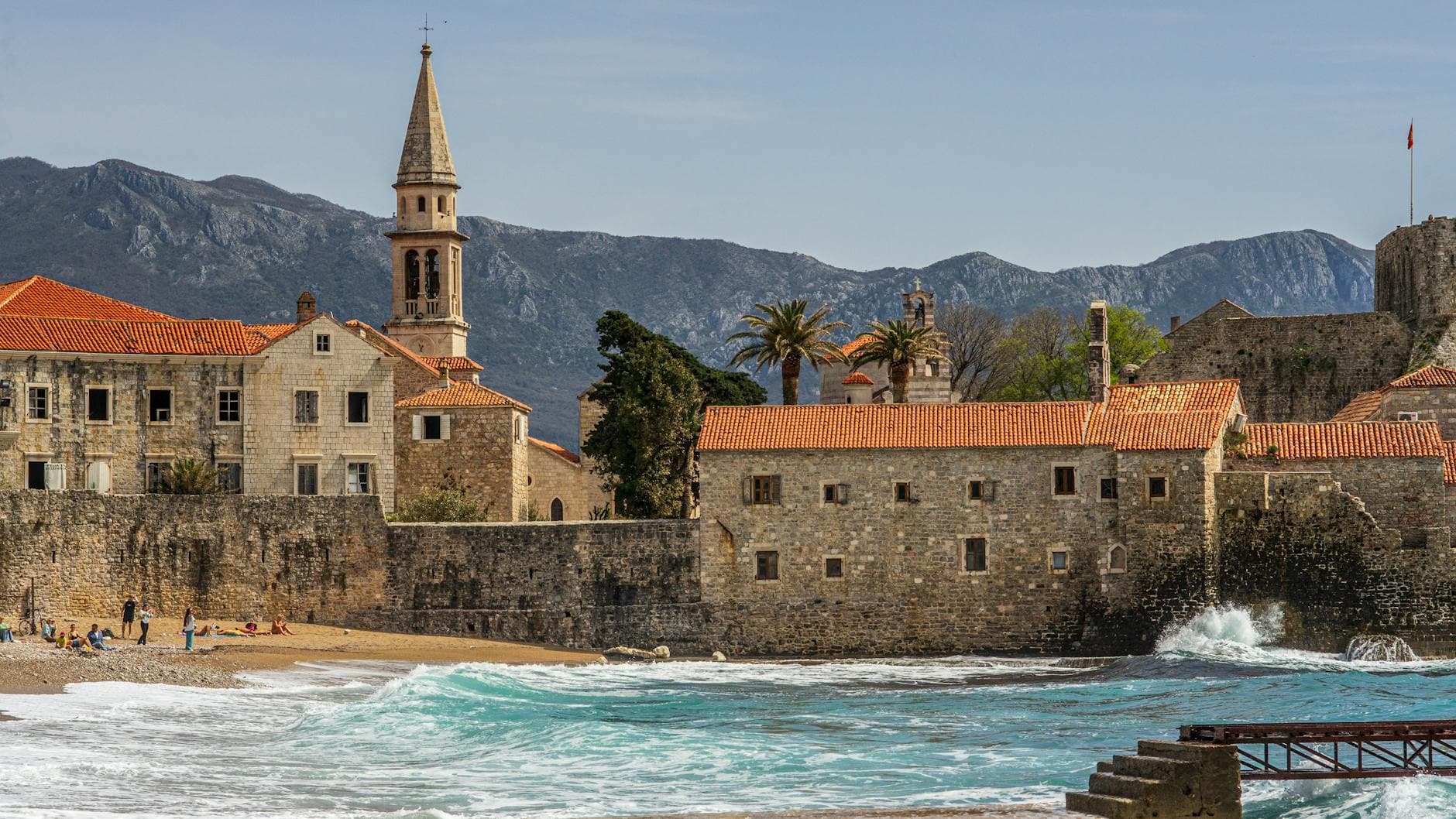 Scenic view of Budva's old town and bell tower by the sea, with mountains in the background.