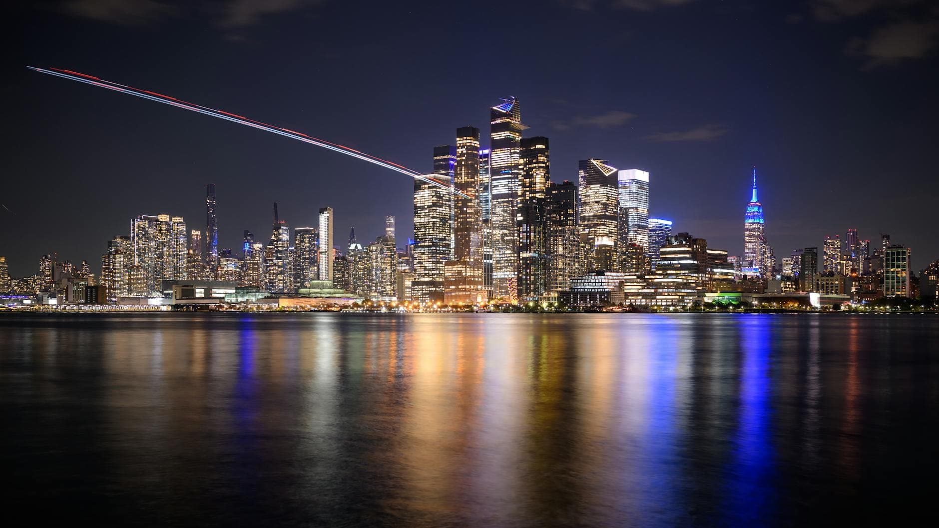 Stunning nighttime view of New York City skyline across the reflective waterfront, featuring illuminated skyscrapers.