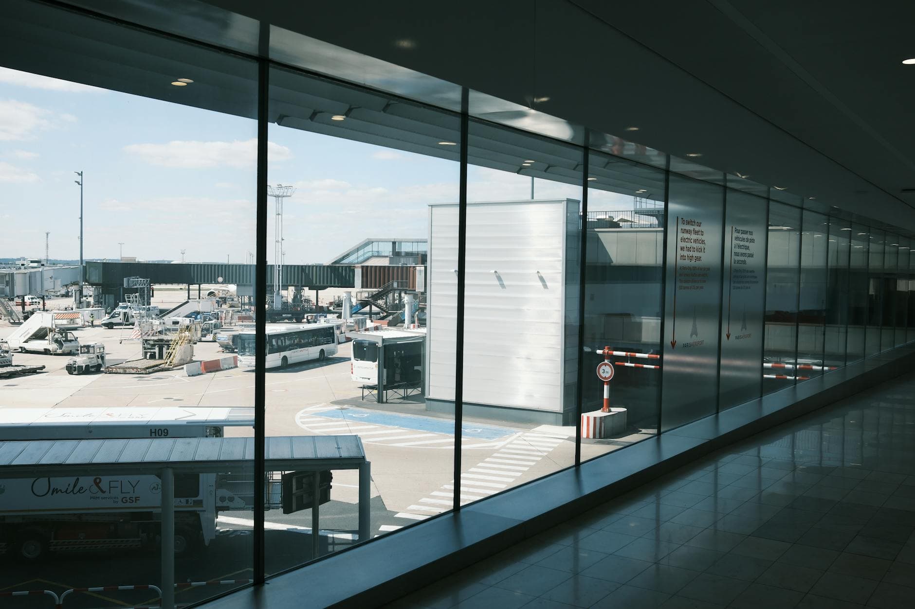 Wide view of an airport terminal hallway and tarmac, showcasing airplanes and buses.