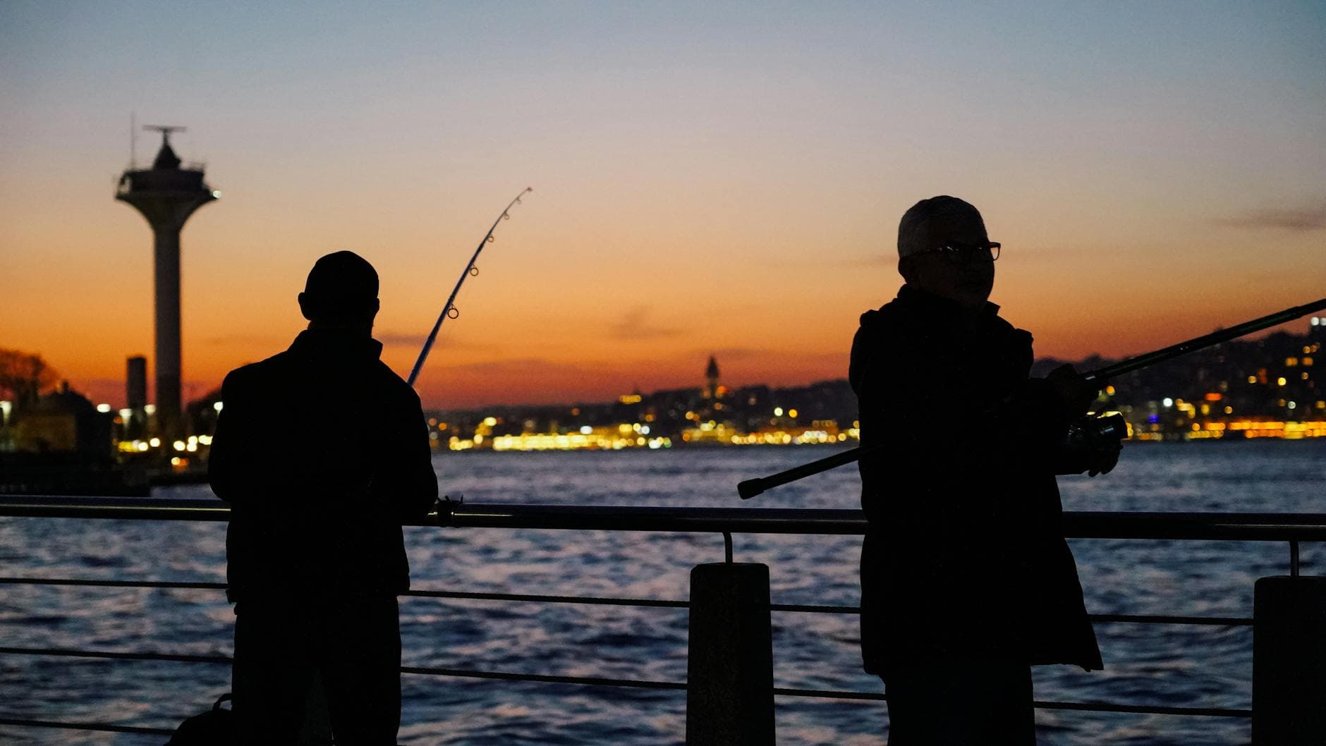 Silhouetted fishermen casting lines at sunset, overlooking the Istanbul skyline in Üsküdar, Turkey.