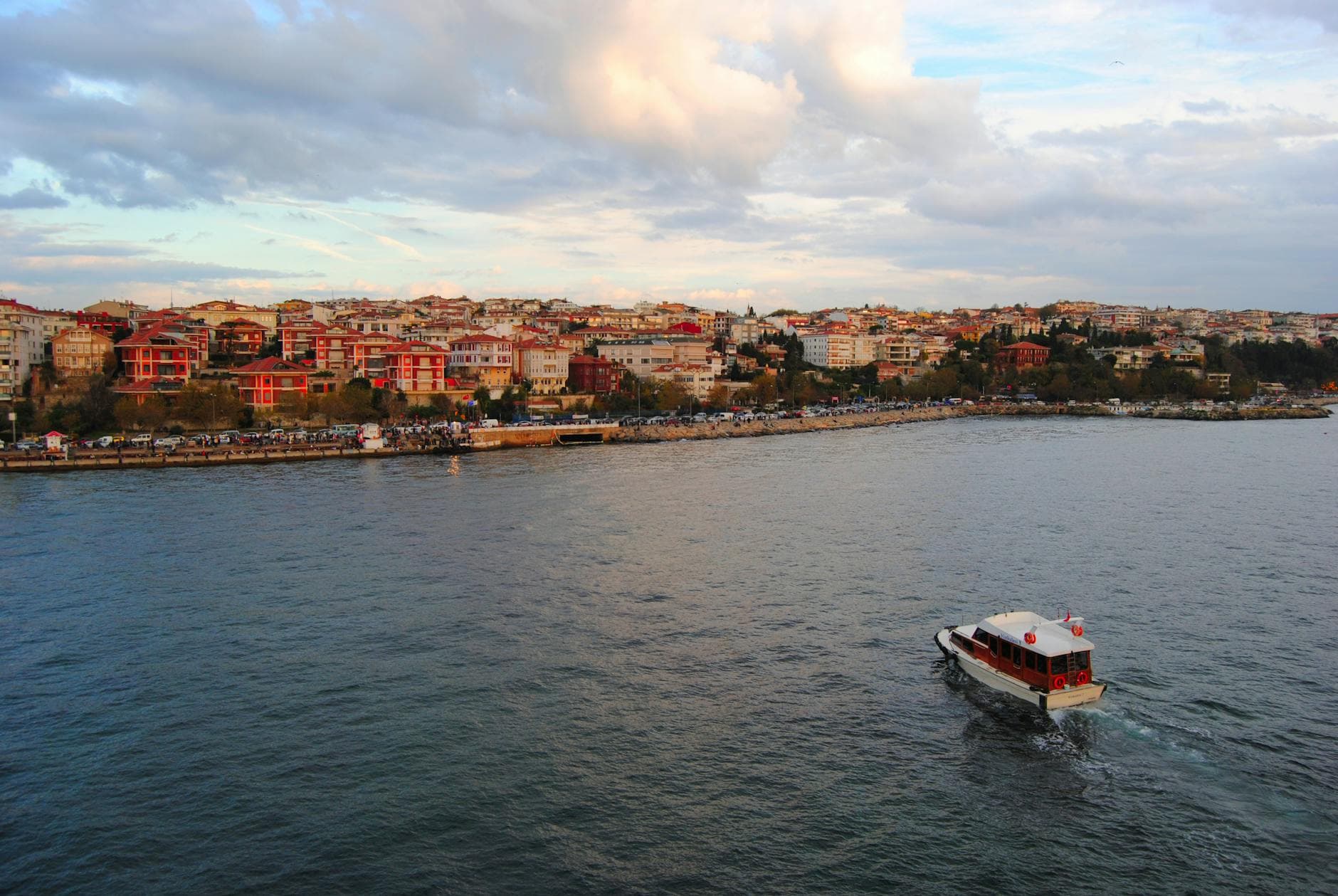 Scenic view of a small boat sailing along the Üsküdar coastline with colorful buildings and a cloudy sky.