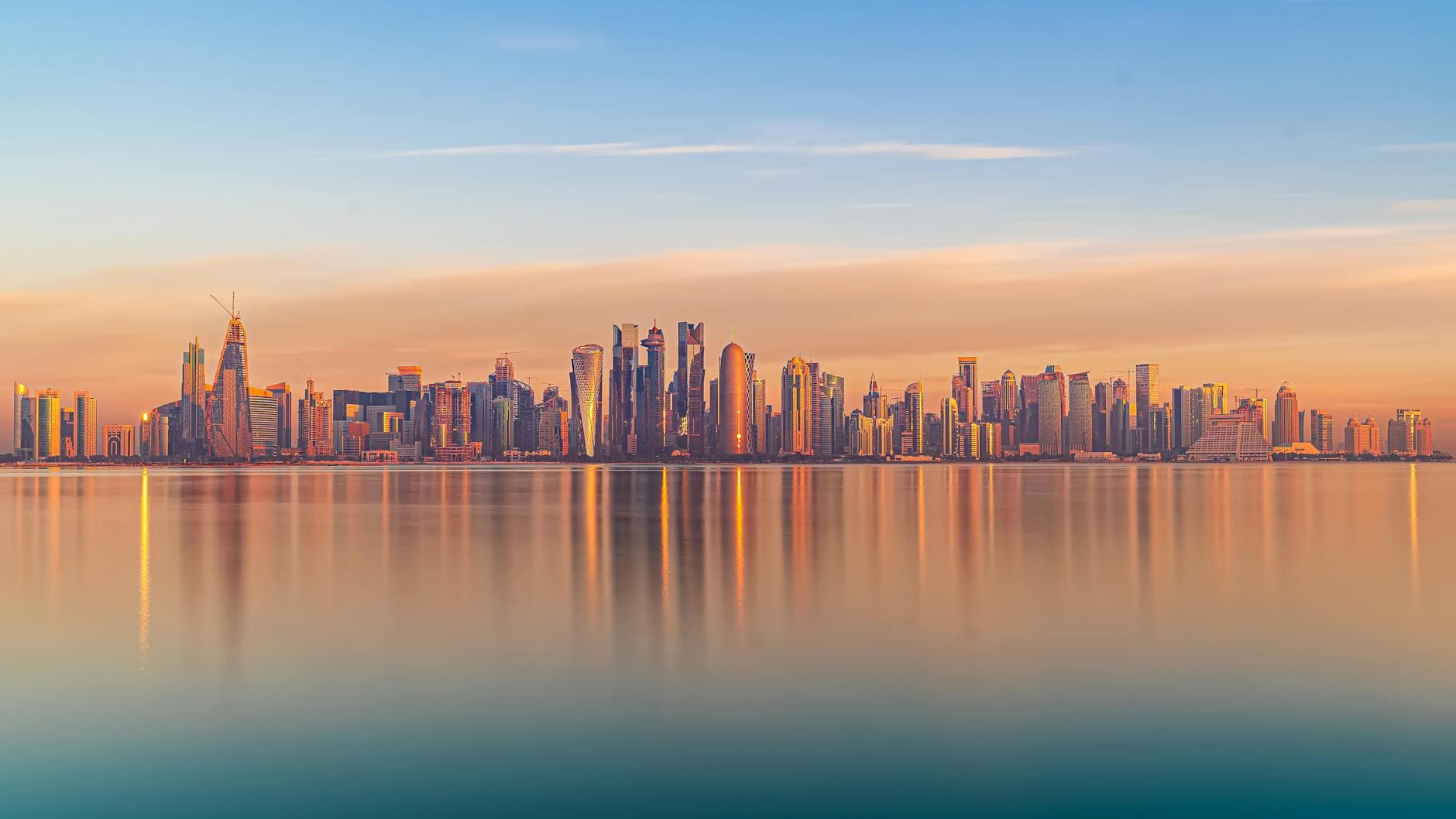 Stunning view of Doha skyline with skyscrapers reflecting in calm water under a sunset sky.