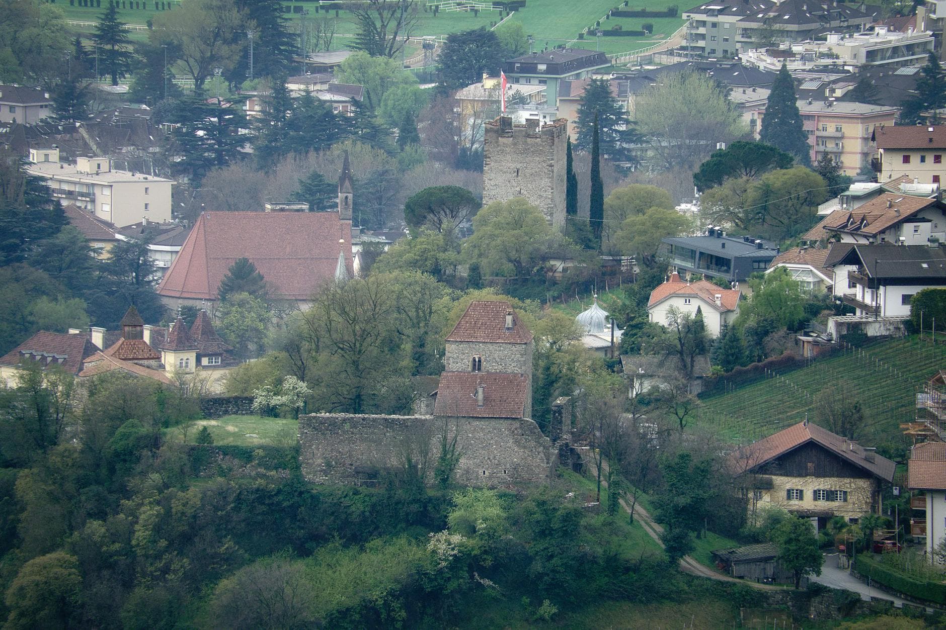A picturesque view of a historic castle in Merano, Italy, surrounded by lush spring greenery and a quaint townscape.