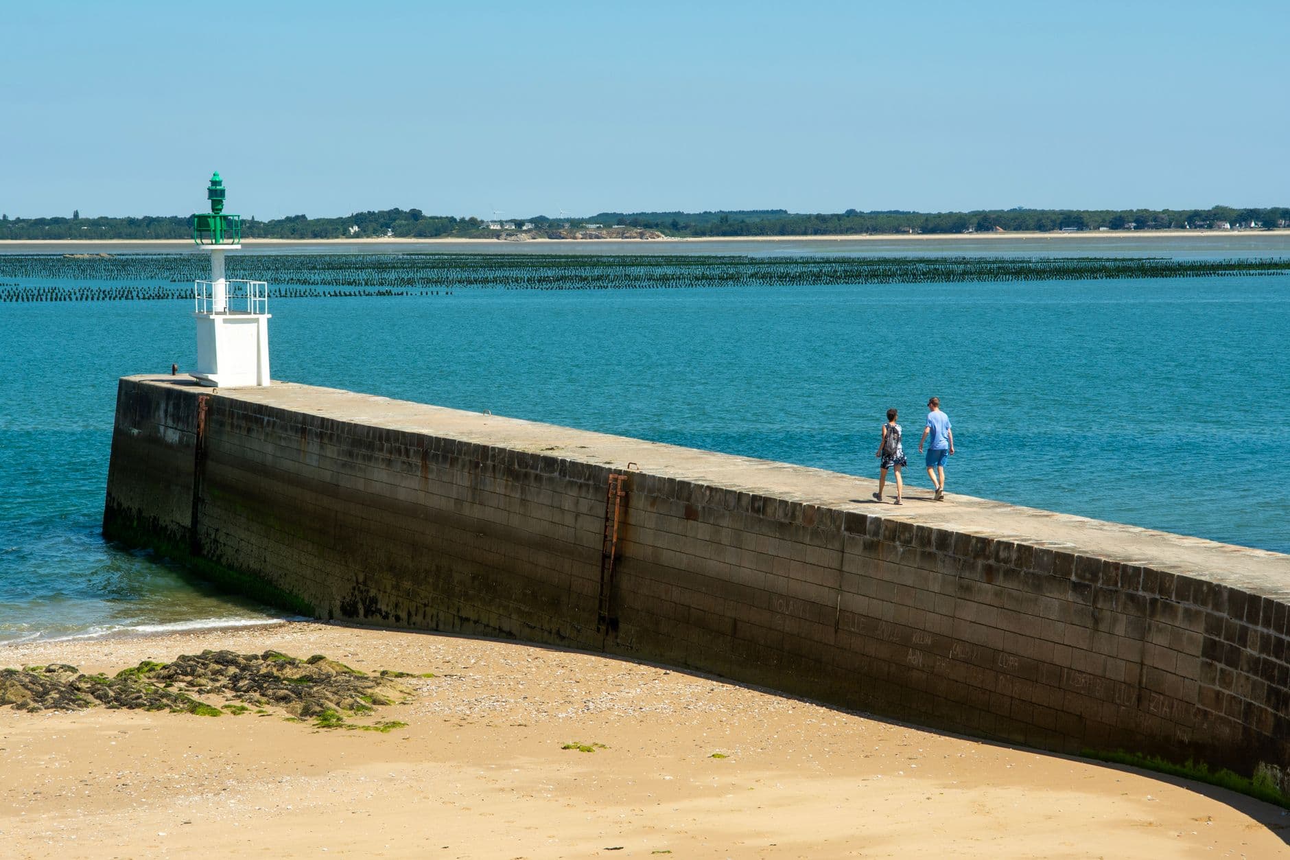 A tranquil scene with a lighthouse pier, beach, and clear blue ocean at Pointe de Merquel, ideal for summer relaxation.