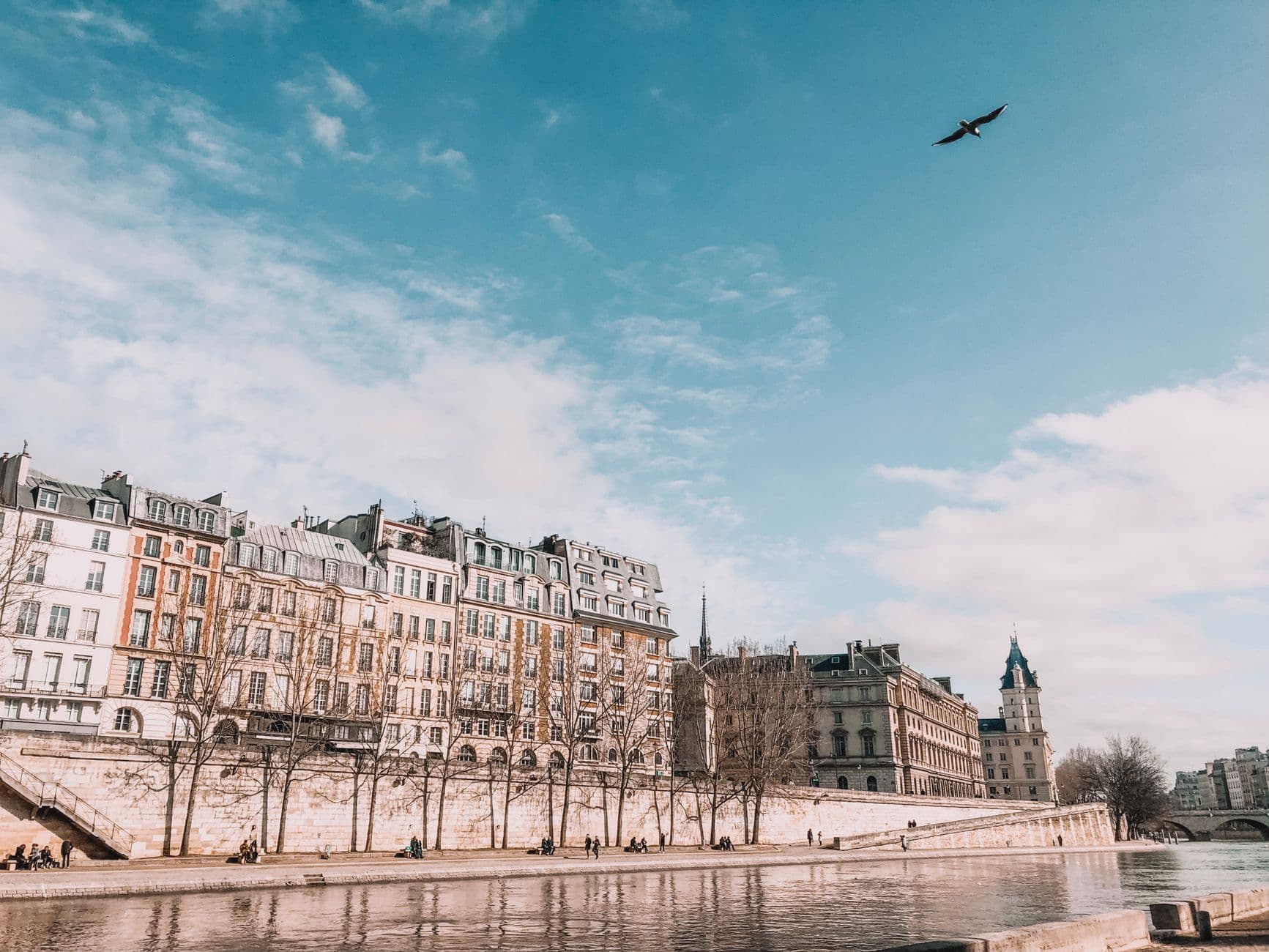A scenic view of historic buildings lining the river under a blue sky, with people strolling along the waterfront.