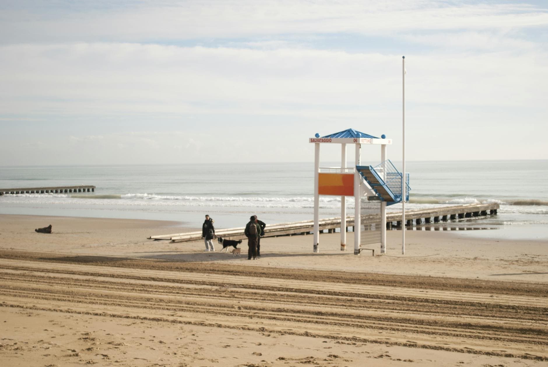 A tranquil beach scene in Caorle, Italy, featuring people walking their dog by a colorful lifeguard tower.