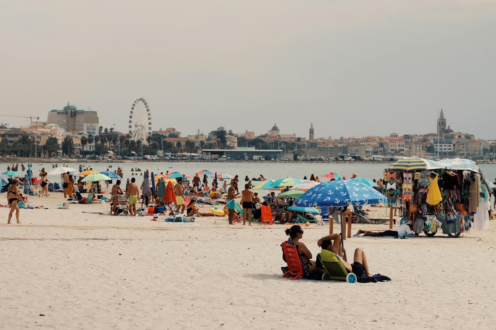 Vibrant beach scene in Alghero, Sardinia with people, umbrellas, and city skyline in the background.