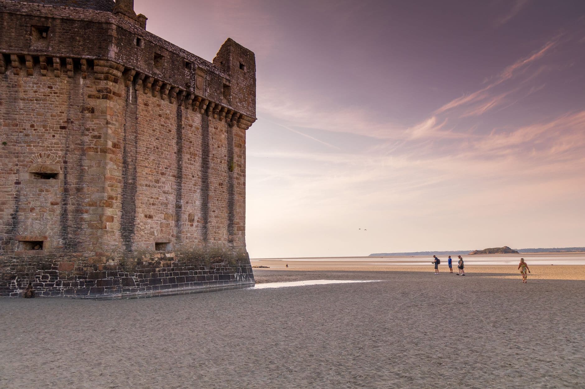 Scenic view of Le Mont-Saint-Michel's ancient fortress with people walking on the beach at sunset.